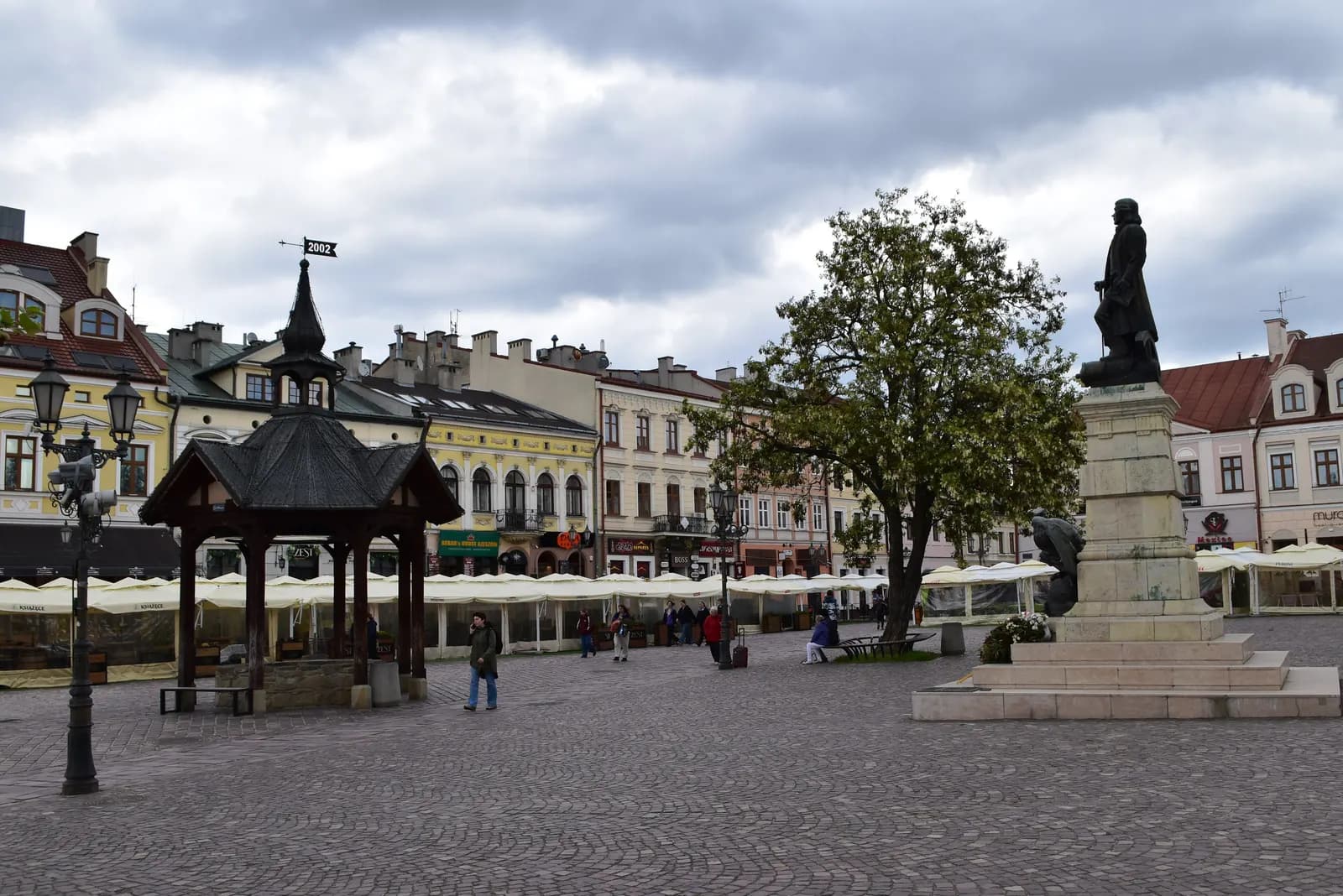 The Market Square in Rzeszów with a wooden roofed pavilion, a tree, a monument on a tall plinth and terraces in front of the houses