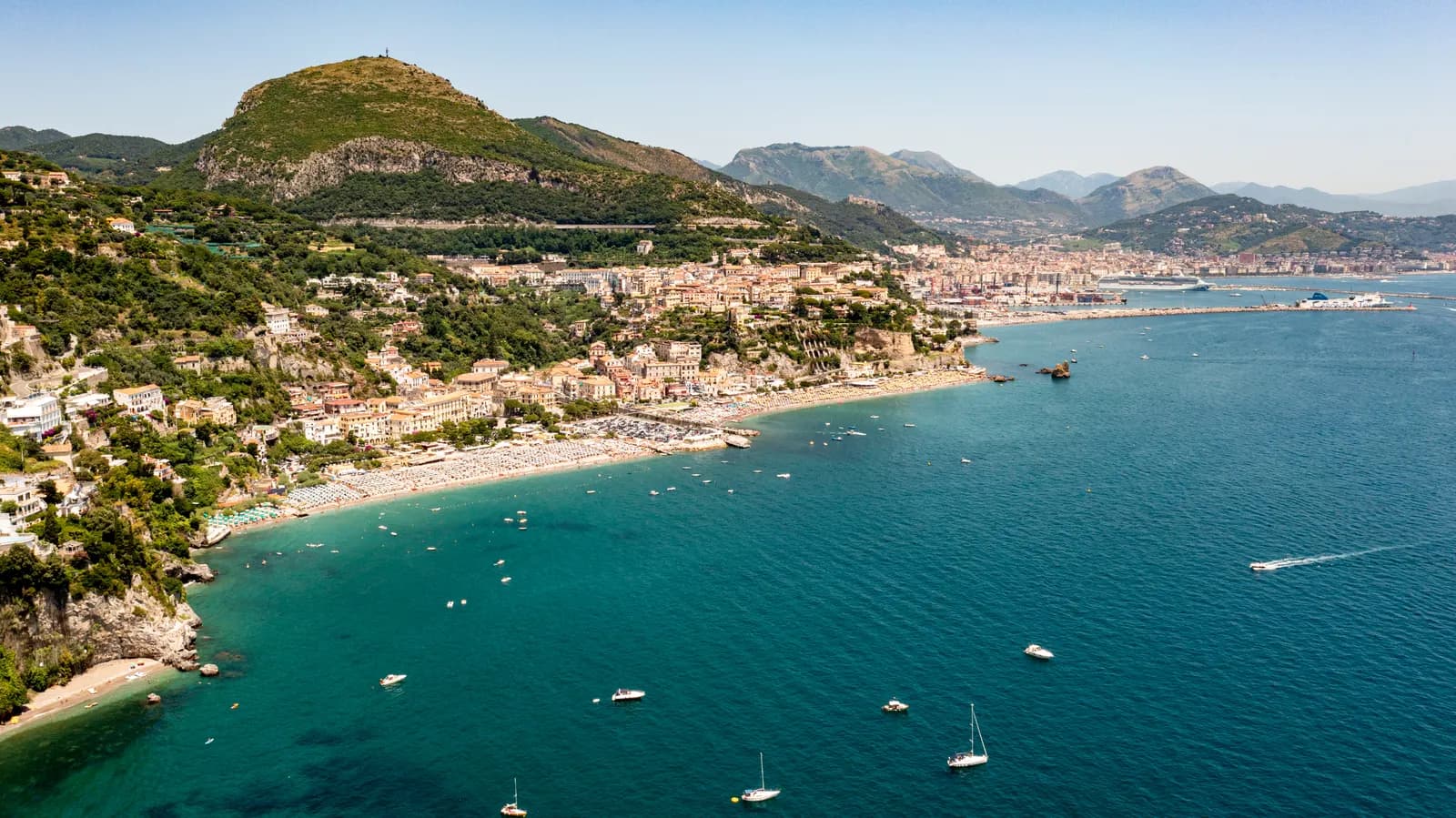 The Gulf of Salerno from above, with a crowded beach and a hillside town in the foreground and the long breakwater and quays of Salerno's port along the shore beyond.
