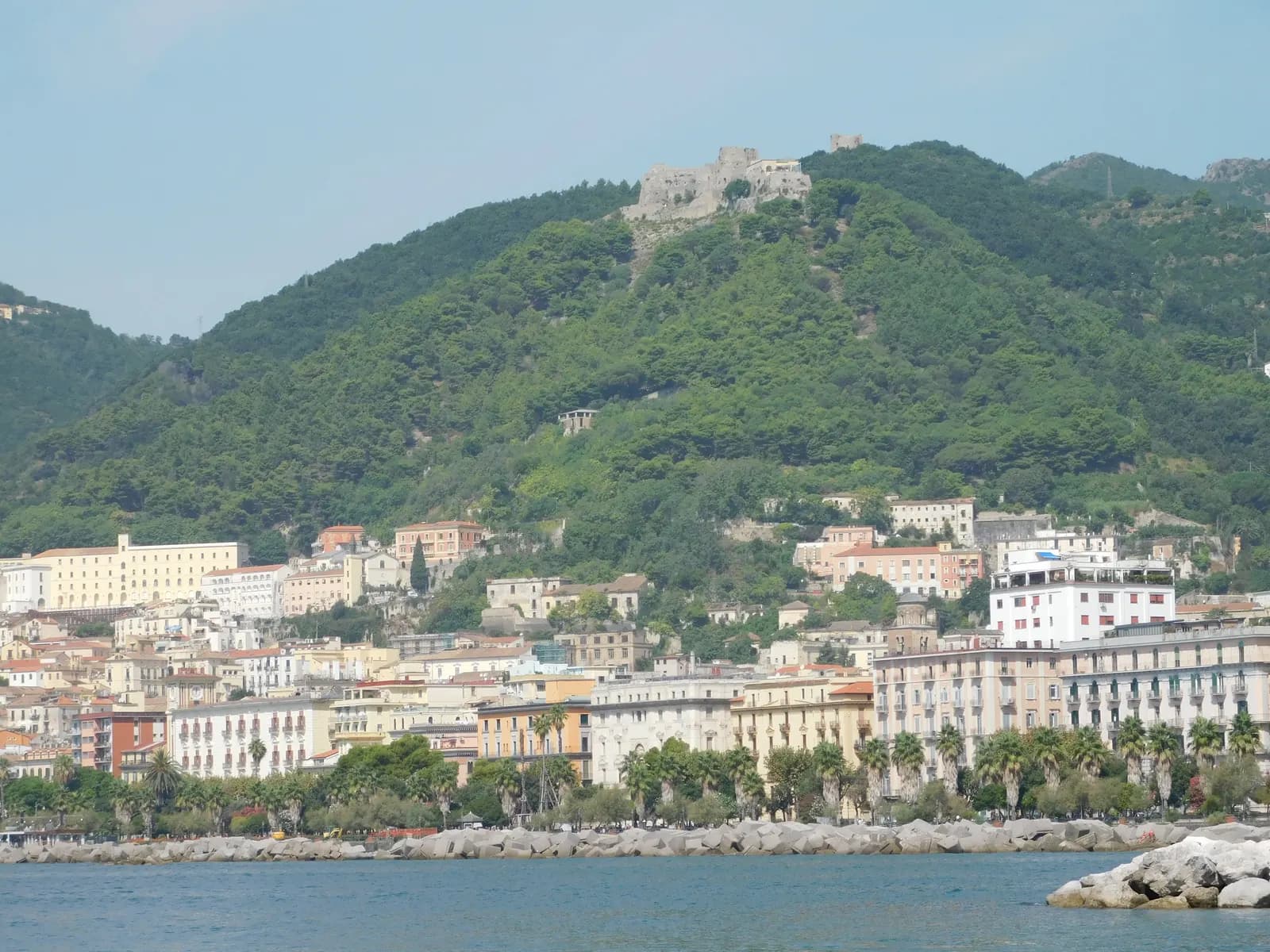 Salerno's seafront seen from the water, with palm trees along Lungomare Trieste, pale apartment blocks behind them and the ruined Castello di Arechi on the wooded hill above.