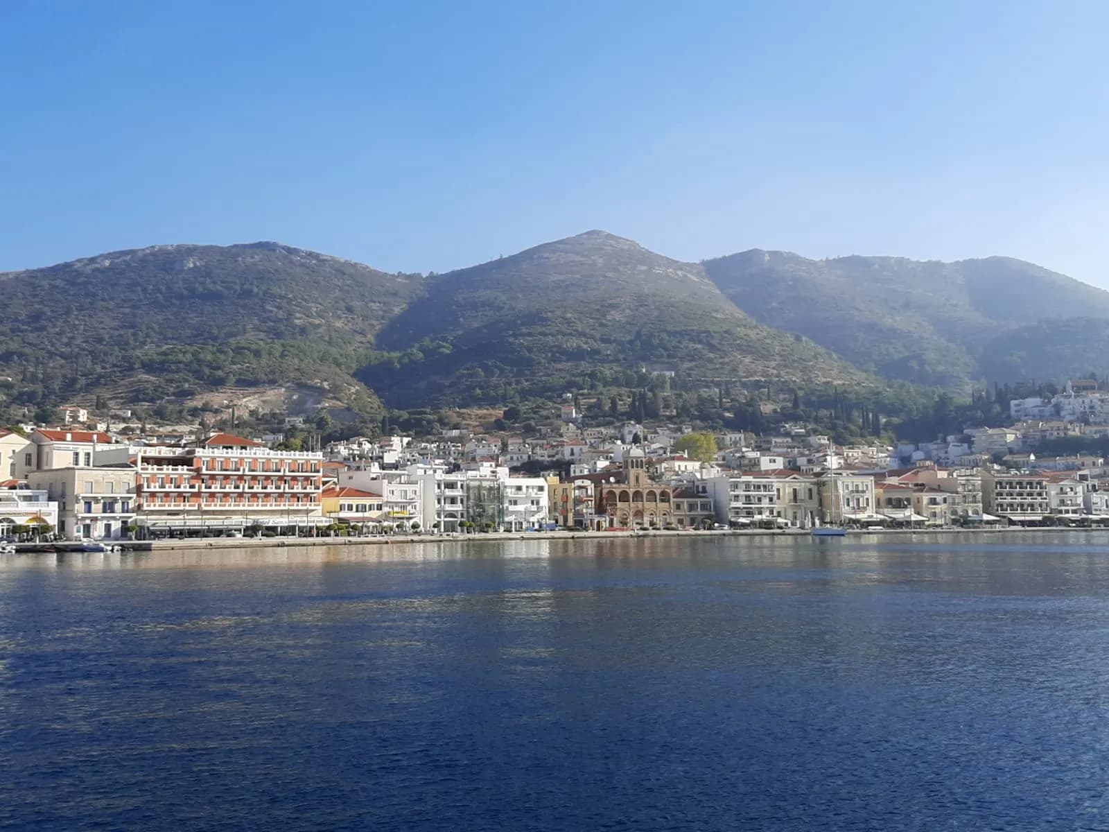 The waterfront of Samos town seen from the water, with hotels and houses along the quay below green hills