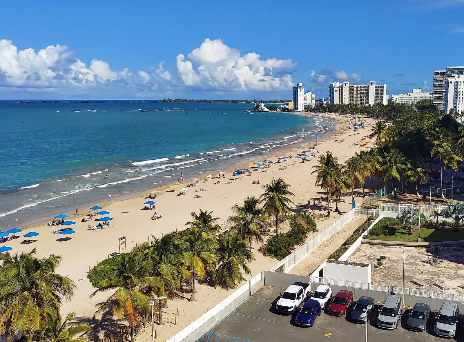 Isla Verde beach seen from above, with blue umbrellas on the sand, palm trees, hotel towers along the shore and a car park in front