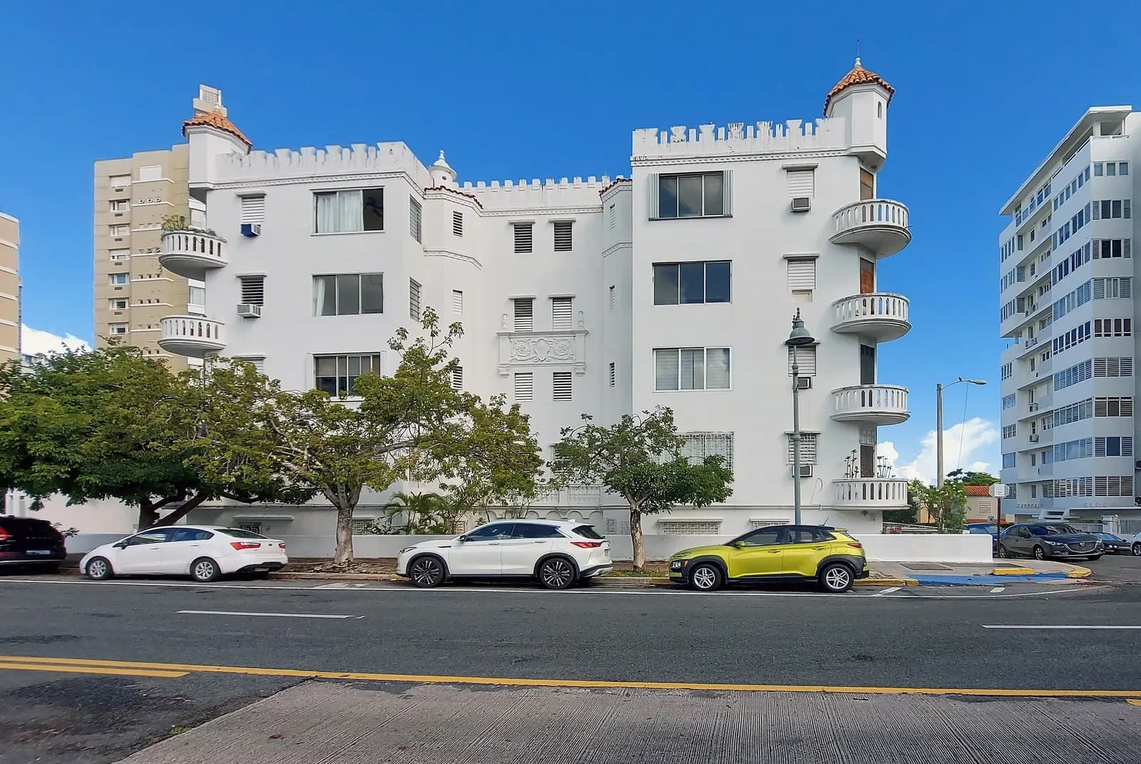 The white Alcazar apartment building in Miramar, with small towers and rounded balconies, parked cars and taller condominium towers beside it