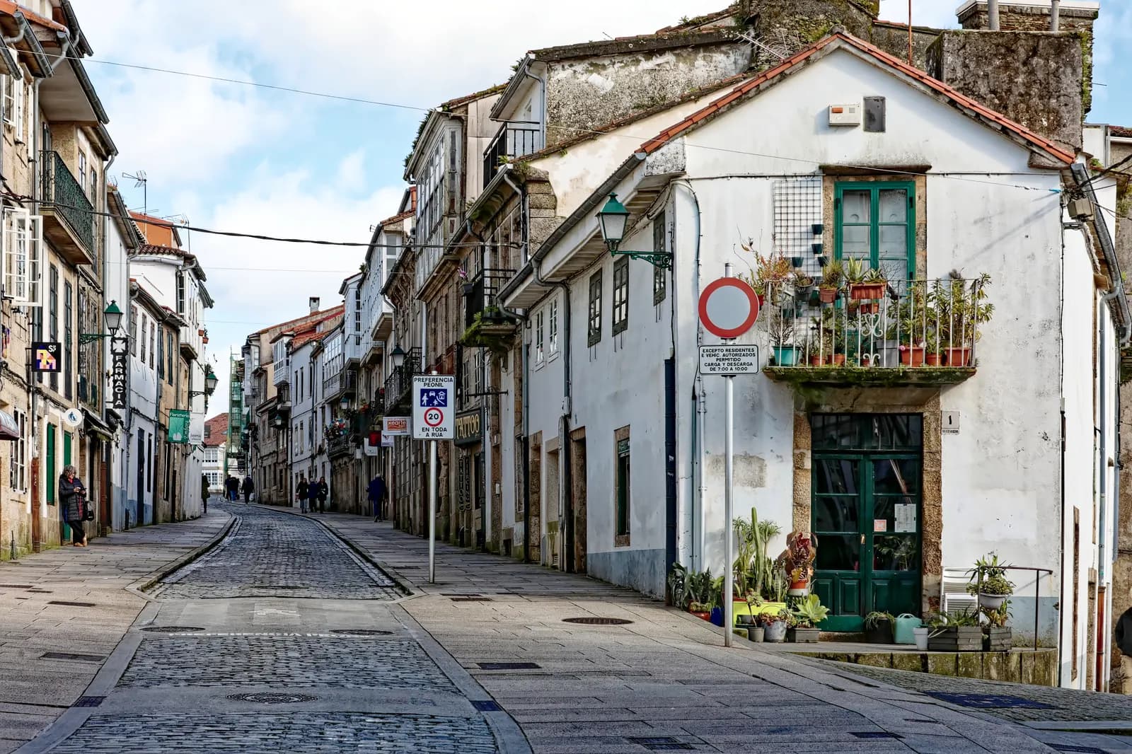 Rúa de San Pedro in Santiago, a cobbled street of whitewashed stone houses with balconies and potted plants, and traffic signs at the corner.