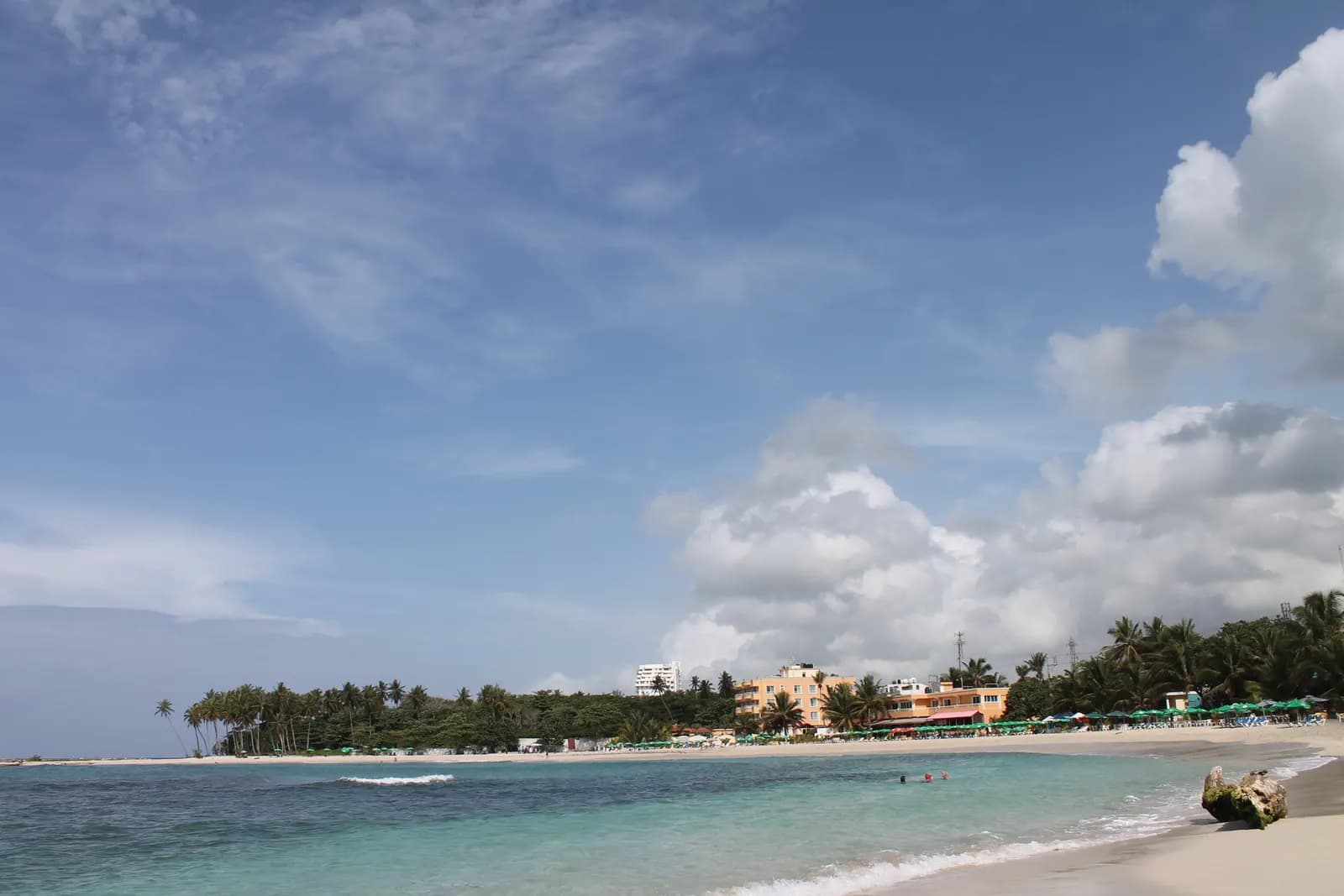 The public beach at Juan Dolio, with shallow turquoise water, sand, palm trees and low buildings along the shore