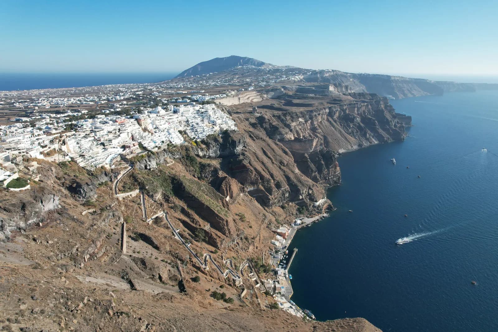 Aerial view of Fira spread along the top of the caldera cliffs, with the zigzag path down to the old port, the sea on the right and a mountain in the distance