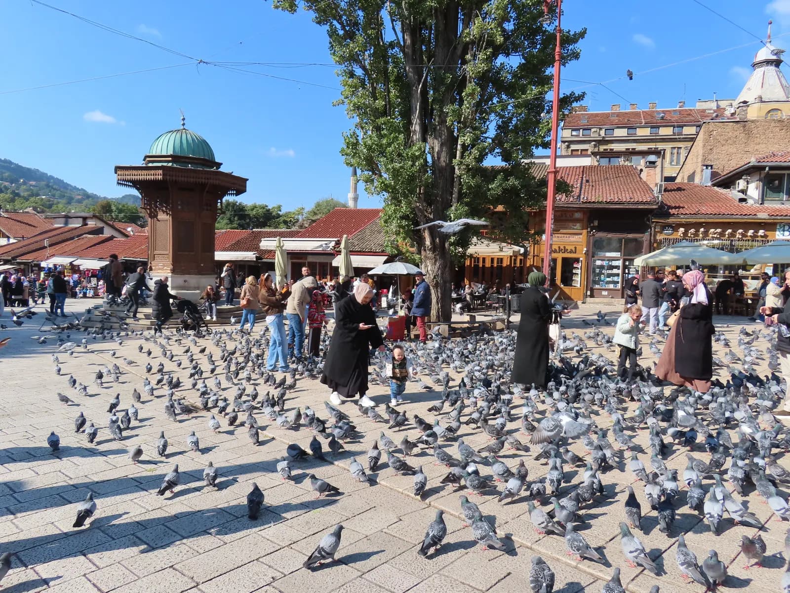 The wooden Sebilj fountain on Baščaršija square in Sarajevo, with pigeons on the paving, visitors and the low roofs of the bazaar behind