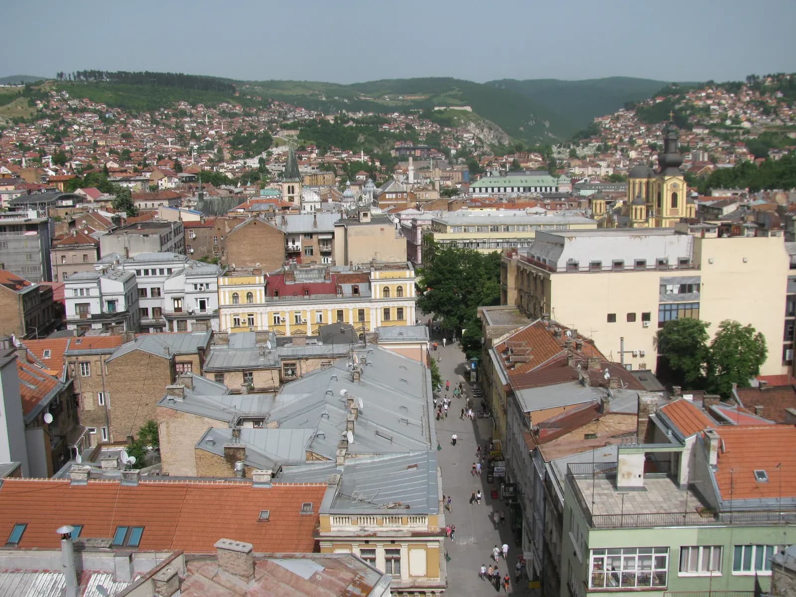 Rooftops of central Sarajevo seen from above, with the Ferhadija promenade running between Austro-Hungarian buildings and the hills behind