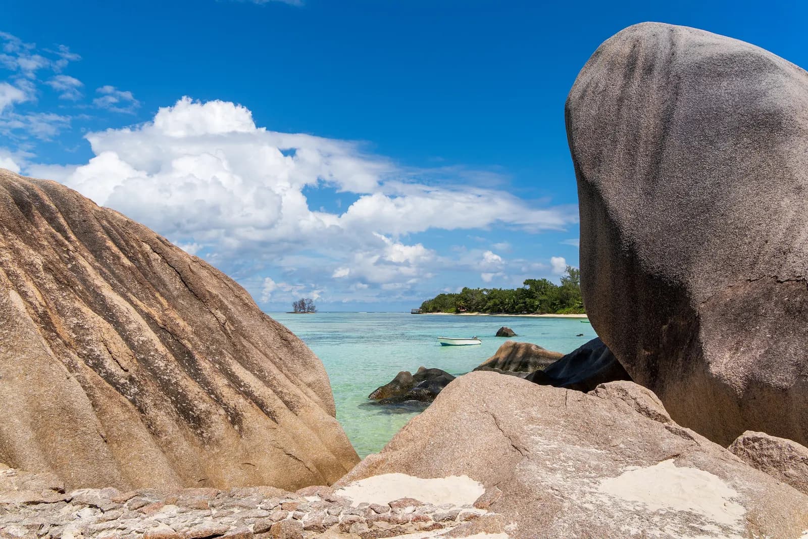 Granite boulders framing the shallow turquoise lagoon at Anse Source d'Argent on La Digue, with a small boat and a wooded point under a blue sky