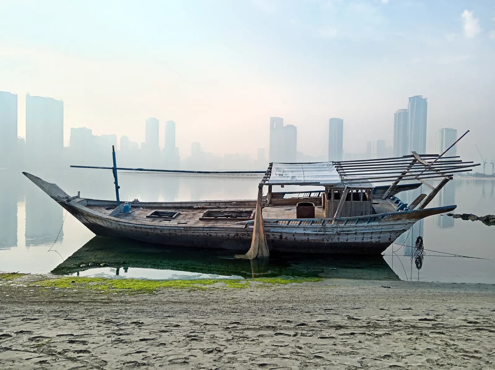 An old wooden boat resting in shallow water off the sand at Al Khan, with tower blocks faint in the morning haze across the water