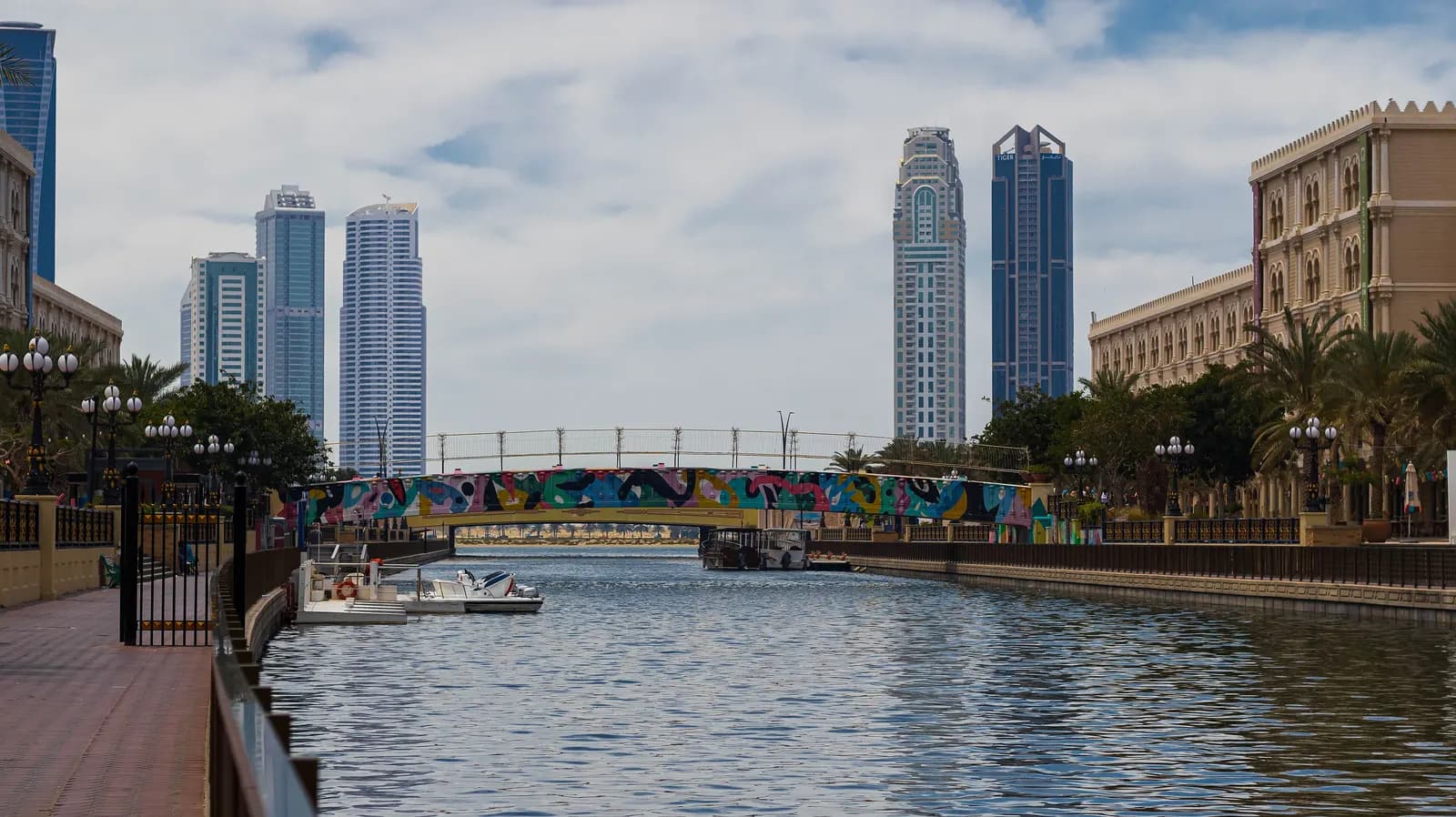 The Al Qasba canal in Sharjah with moored boats, a painted footbridge, palm trees and a promenade, and tower blocks behind