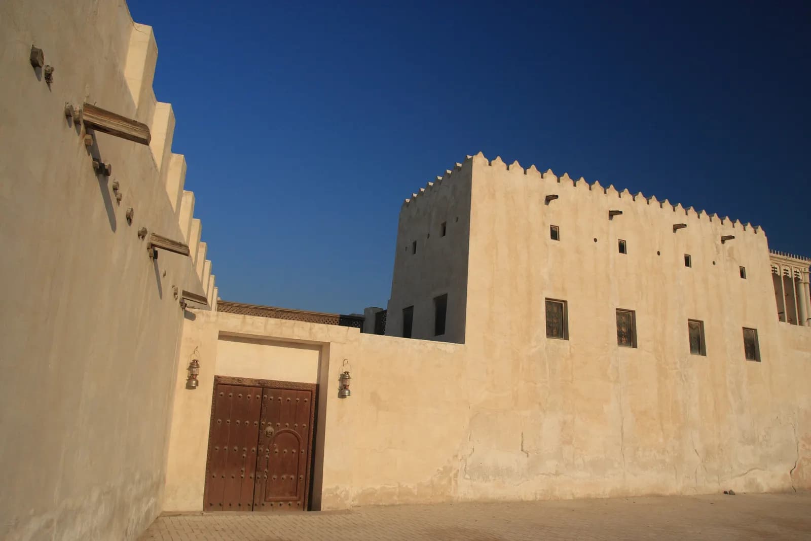 Sand-coloured walls with a carved wooden door and a crenellated tower of a restored building in the Sharjah Heritage Area, under a clear blue sky