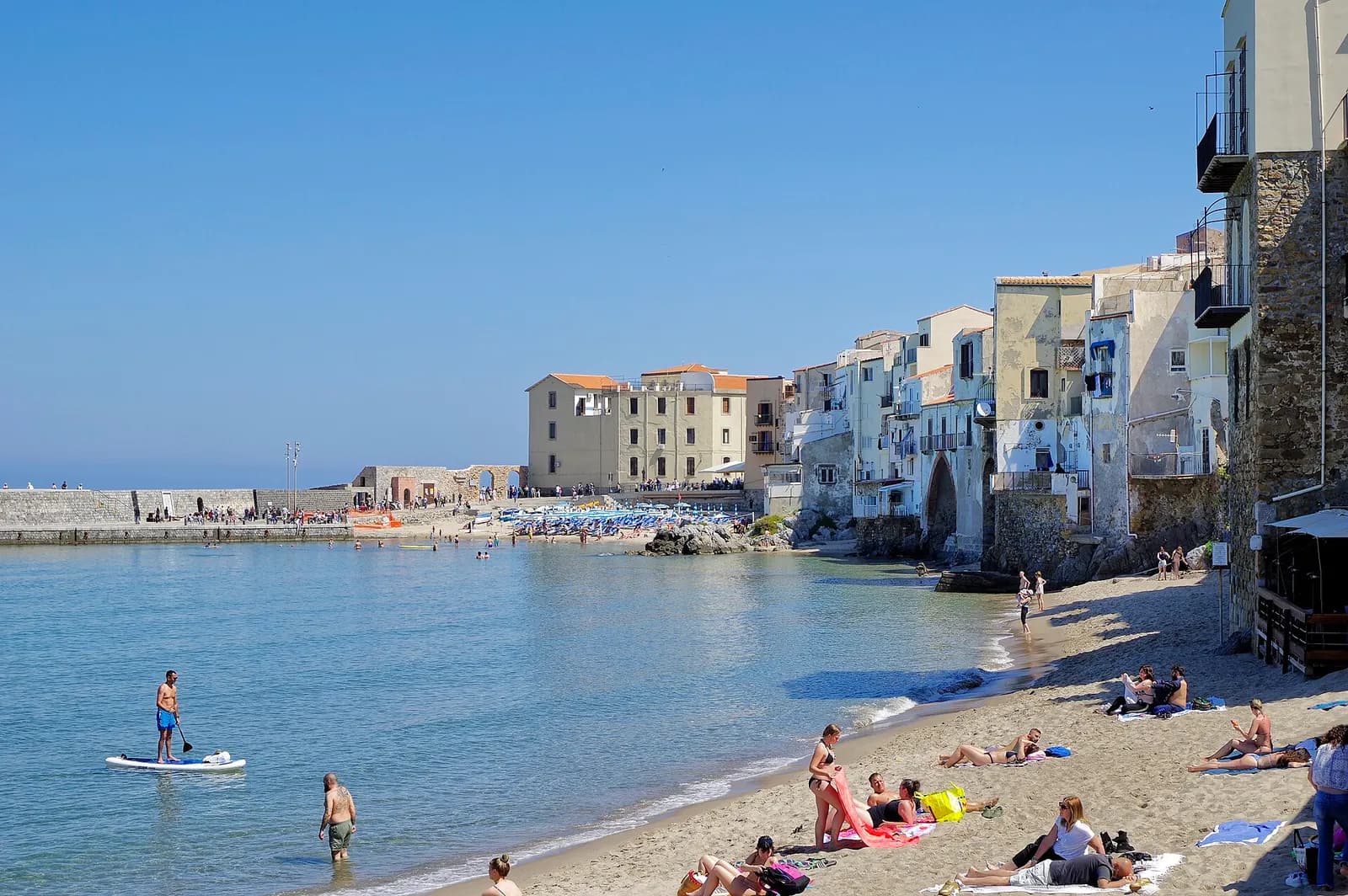 The town beach at Cefalù below old houses built on the sea wall, with swimmers, a paddle boarder and the old harbour wall behind