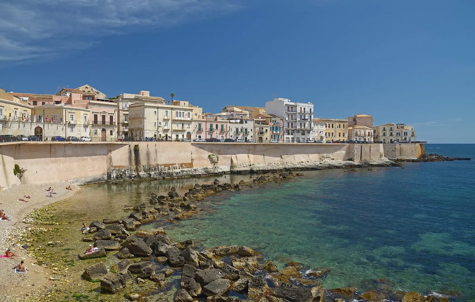 Houses along the Lungomare di Levante in Ortigia above the old sea wall, with clear water, rocks and a small beach below