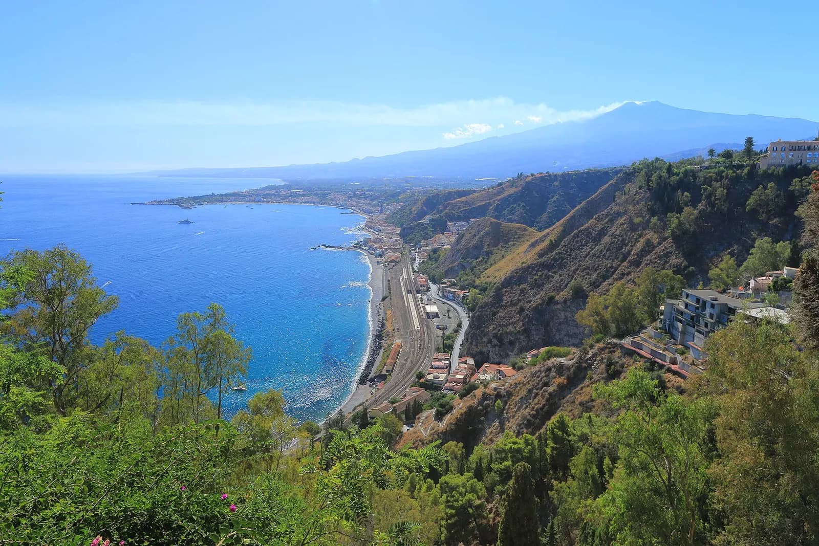 The coast below Taormina curving south to Giardini Naxos, with the railway along the shore and Mount Etna on the horizon