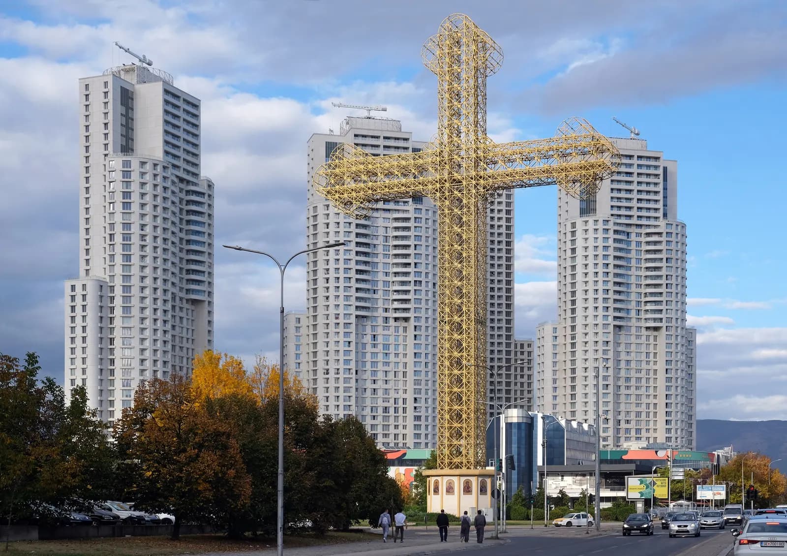 A large golden metal cross in Aerodrom, Skopje, standing in front of a group of white residential towers