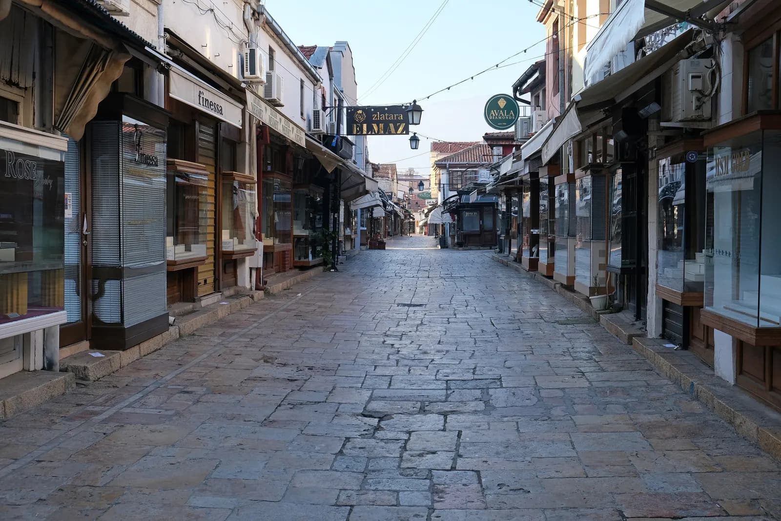 An empty stone-paved lane in the Old Bazaar in Skopje, lined with small shops with wooden shutters and hanging signs