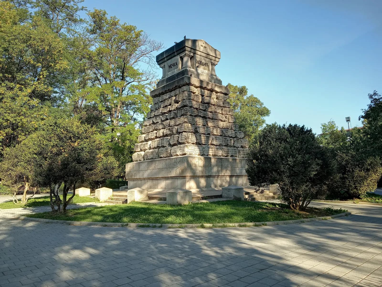 The stone Doctors' Monument in the Doctors' Garden in Sofia, ringed by trees, shrubs and lawn, with a paved path in the morning sun