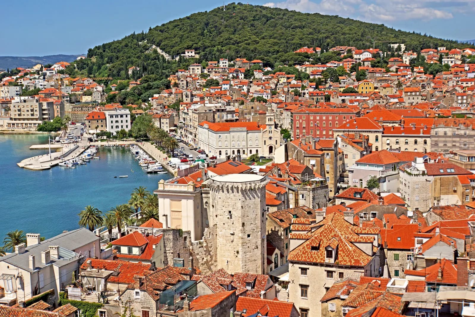 Red roofs of Split old town seen from the cathedral bell tower, with a stone tower, the harbour and the green Marjan hill behind