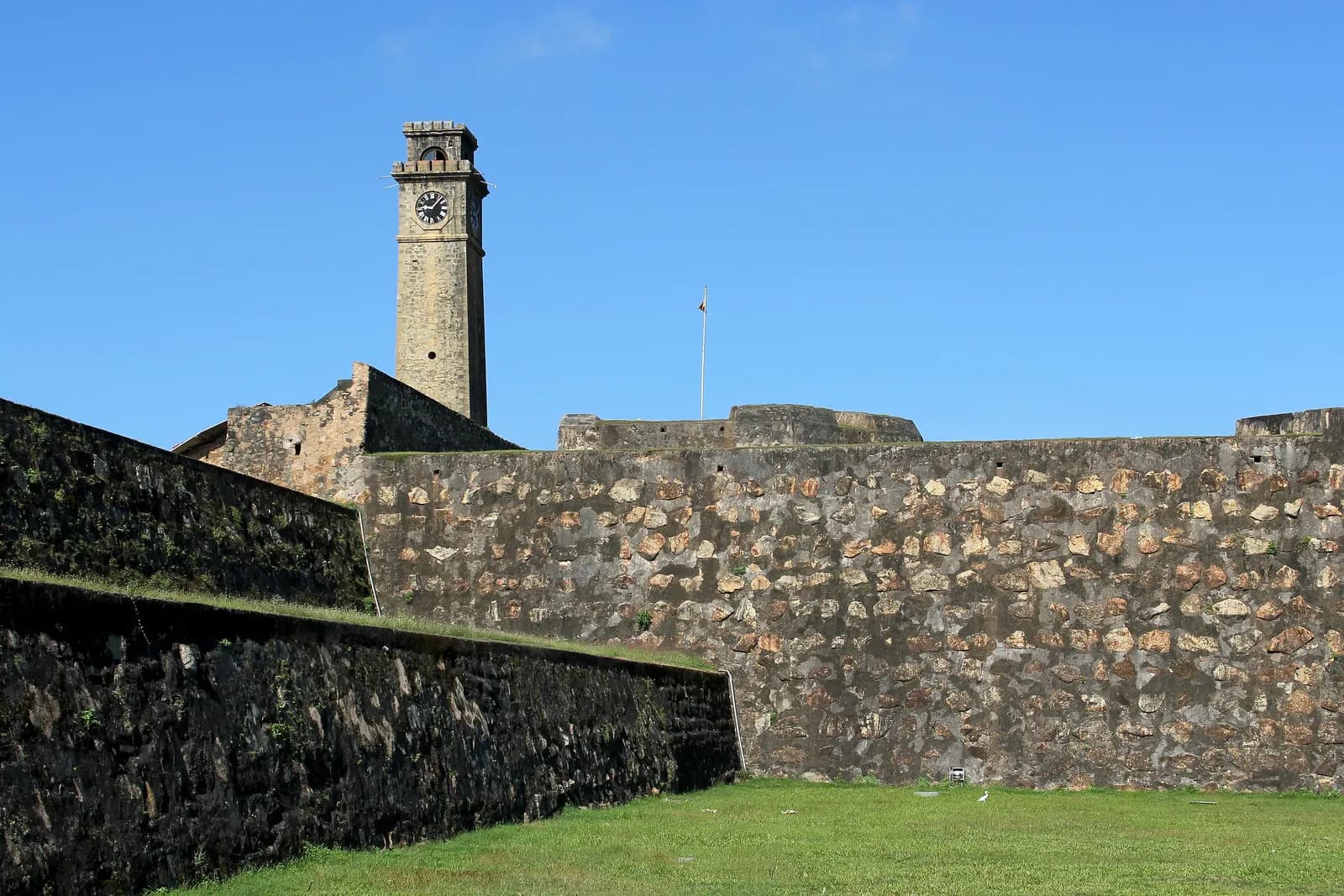 The stone ramparts of Galle Fort with the clock tower rising behind them under a clear blue sky, and a lawn in front