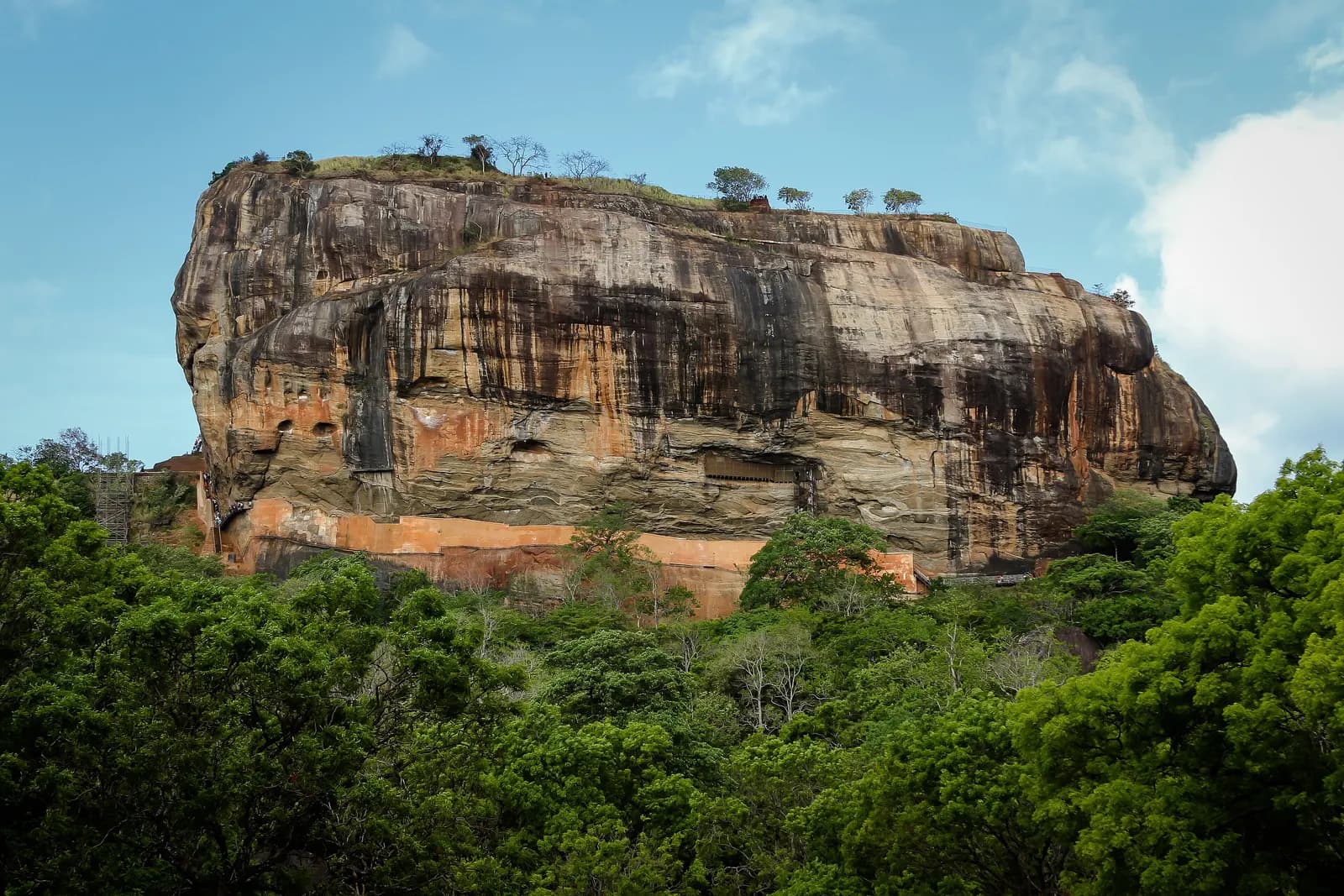 The sheer rock of Sigiriya rising above the forest, with brick walls at its base and trees along the summit