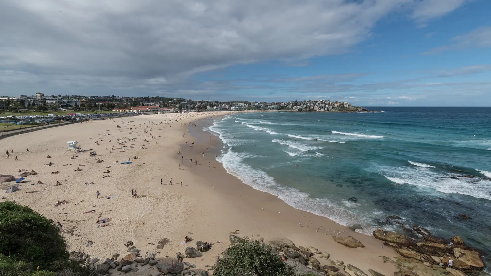 The curve of Bondi Beach with sunbathers on the sand and waves breaking along the shore, and houses and apartment blocks behind the beach and on the far headland