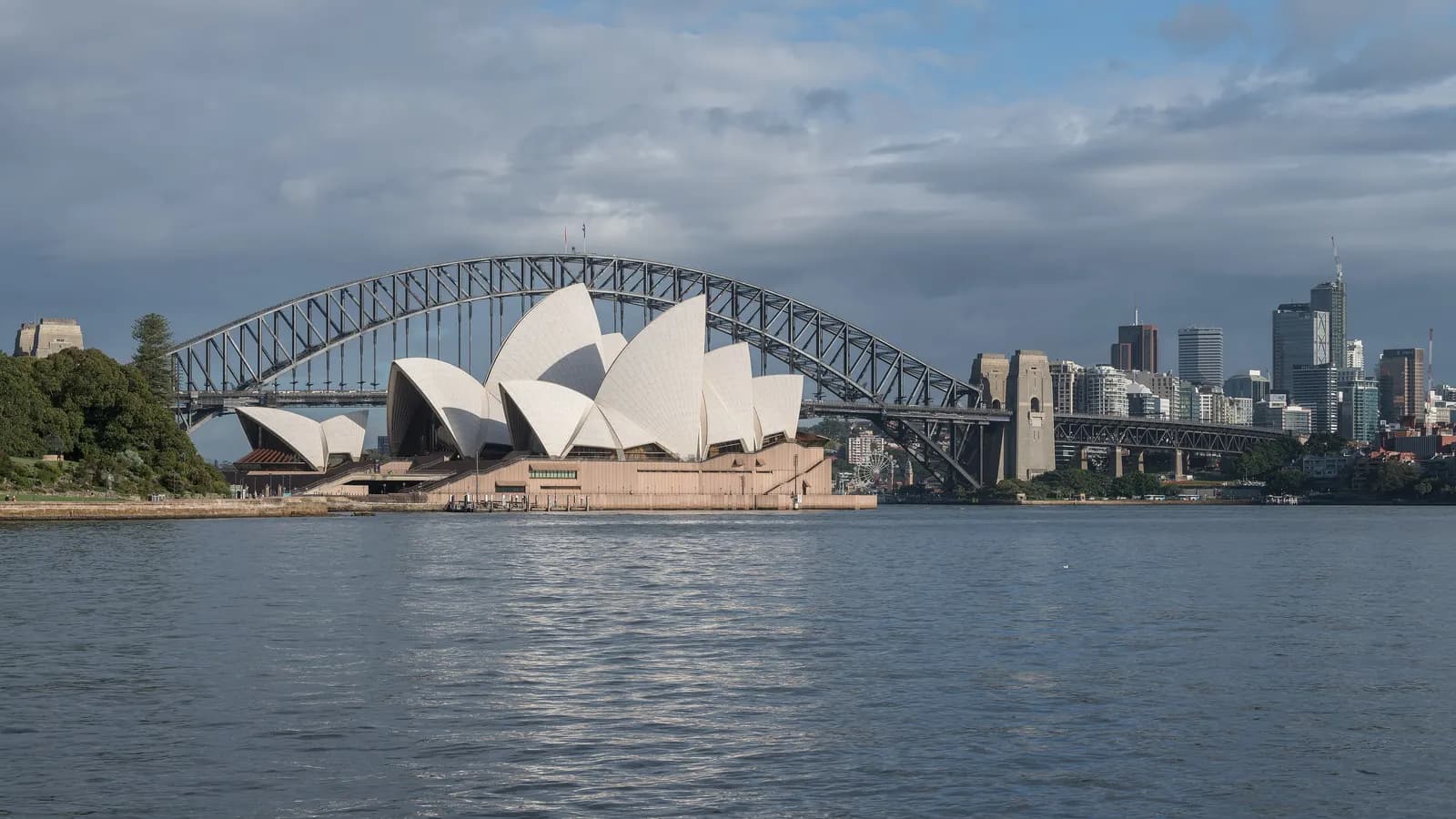 The Sydney Opera House in front of the Sydney Harbour Bridge, seen across the harbour, with office towers on the far shore under a cloudy sky