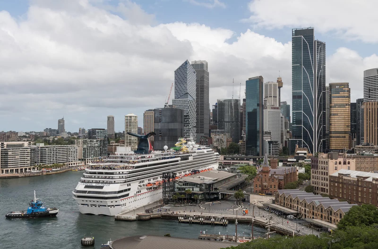 A cruise ship berthed at Circular Quay beside the old sandstone warehouses of The Rocks, with the towers of the Sydney CBD behind, seen from the Harbour Bridge
