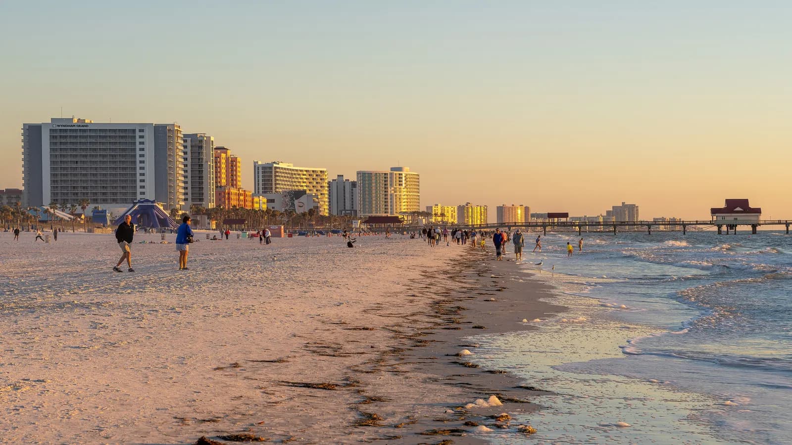 Hotel towers along the wide sandy shore of Clearwater Beach in late afternoon light, with people walking at the water's edge and a pier in the distance