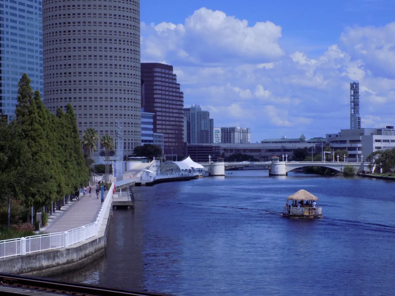 The Tampa Riverwalk with walkers beside the Hillsborough River below a round tower and other downtown buildings, with a thatched party boat on the river and a road bridge beyond