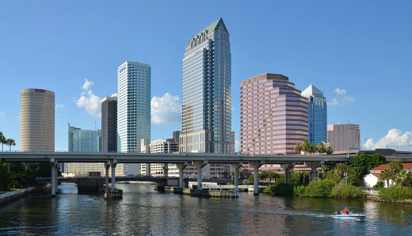 Downtown Tampa office towers above the Hillsborough River, with an elevated road bridge crossing the water and a small motorboat in the foreground under a blue sky