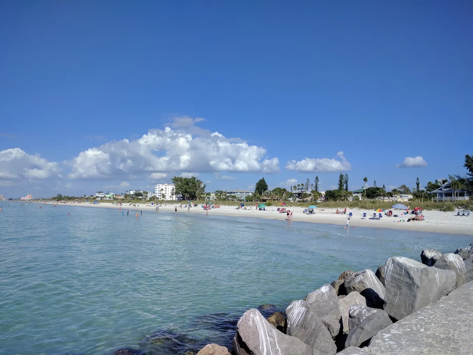 The beach at Pass-a-Grille in St. Pete Beach seen from a rock jetty, with clear shallow water, beachgoers under umbrellas and low houses among the trees