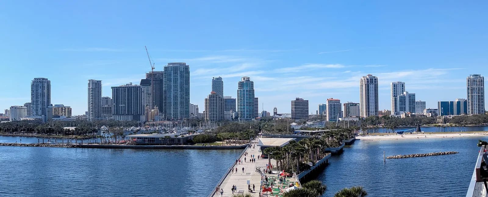 The downtown St. Petersburg skyline seen from the St. Pete Pier, with people walking along the pier, a marina with sailboats and a small sandy beach on the bay