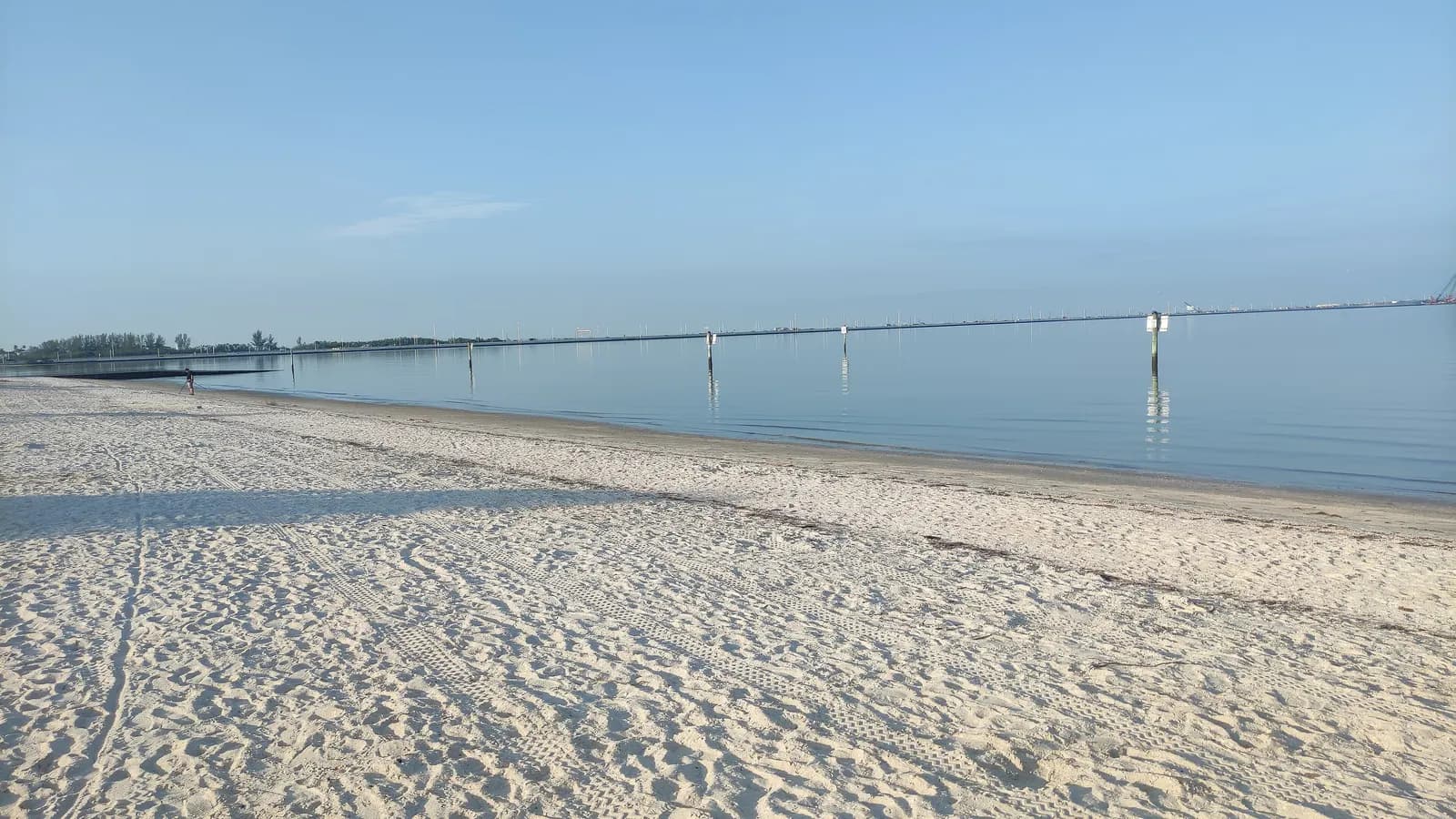 The white sand beach at Cypress Point Park in Tampa, with calm bay water, marker posts and a long low causeway on the horizon under a clear sky