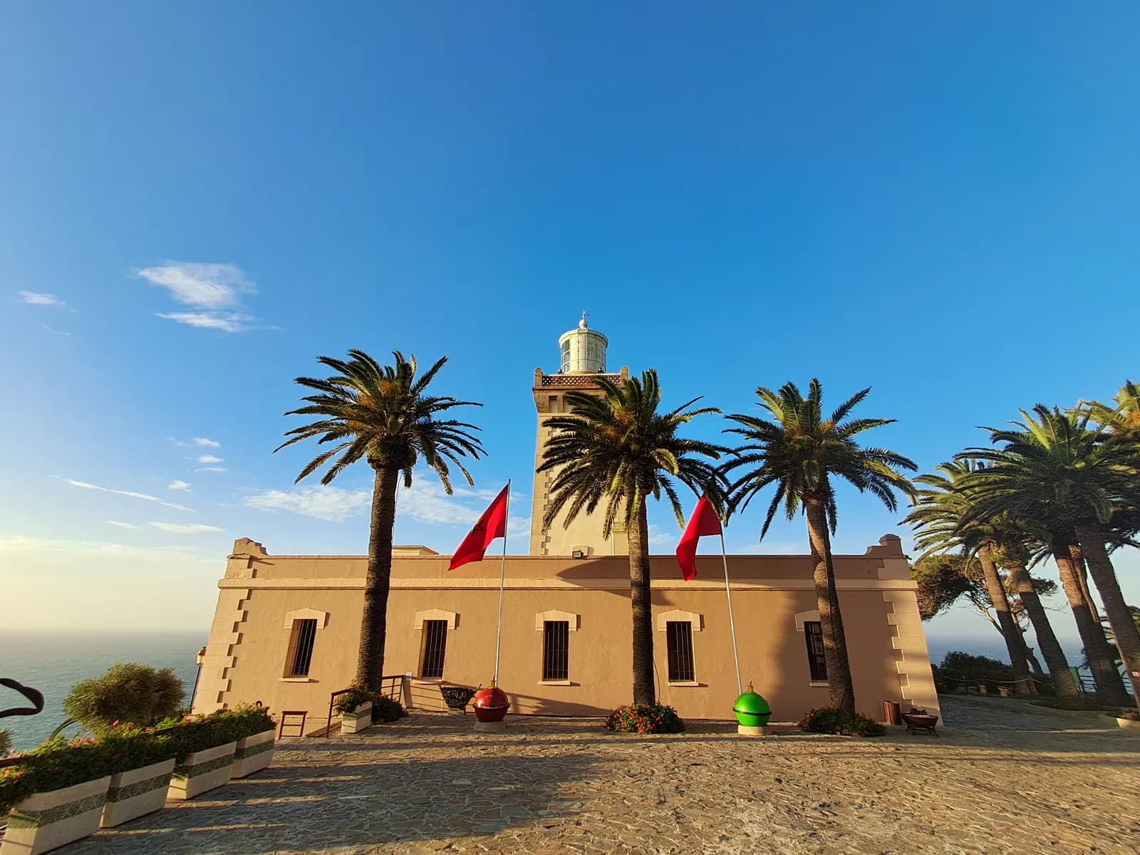 The Cap Spartel lighthouse west of Tangier, a square stone tower above a low sand-coloured building framed by tall palms, with the sea on the left under a blue sky