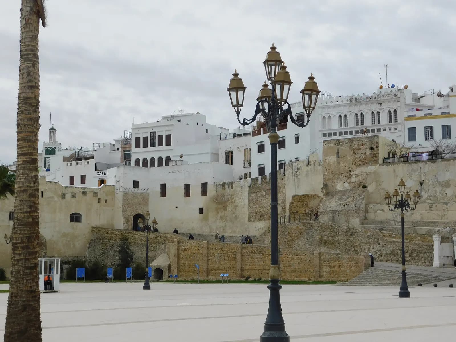 The stone walls of the Kasbah of Tangier under a cloudy sky, with white houses and a green-tiled minaret above them and a wide paved square with lamp posts in front