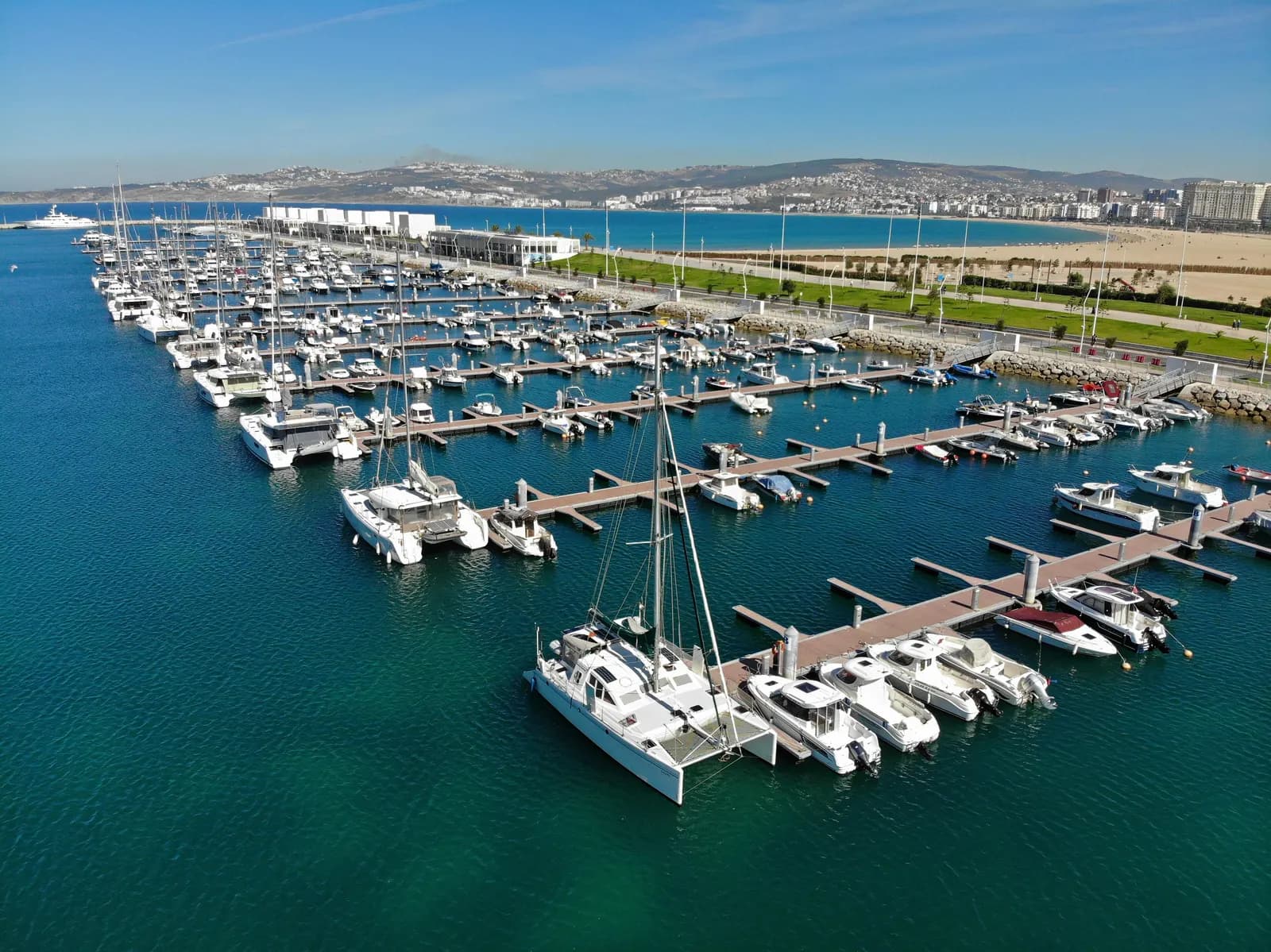 Tanja Marina Bay in Tangier from the air: yachts and catamarans along the pontoons, the seafront promenade and beach behind, and the city along the curve of the bay