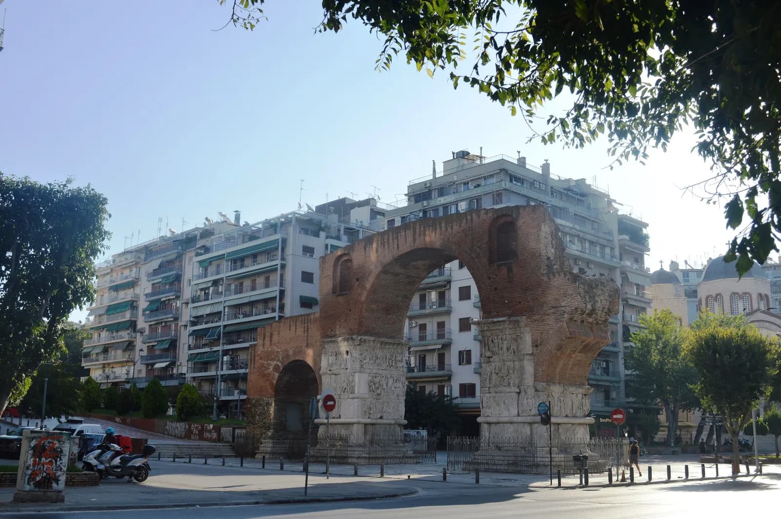 The brick and marble Arch of Galerius, known as Kamara, in central Thessaloniki, framed by trees and apartment blocks in morning light
