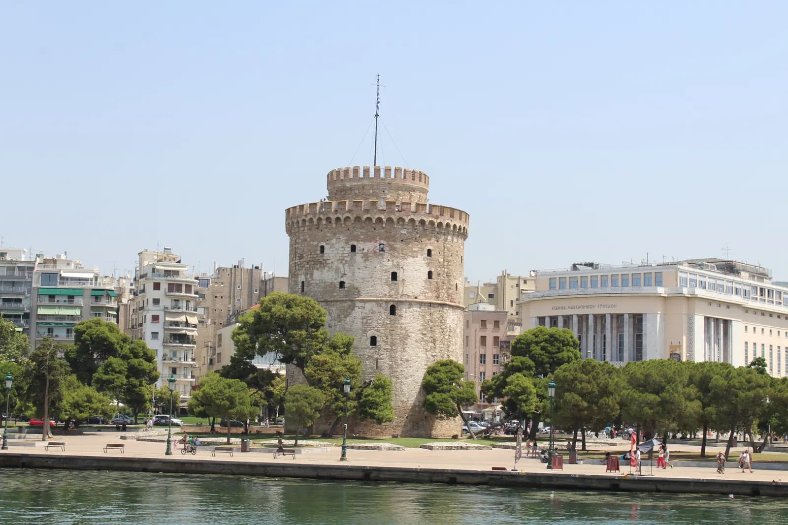 The White Tower of Thessaloniki among pine trees on the waterfront promenade, seen from the sea, with apartment buildings on the left and the Society for Macedonian Studies building on the right