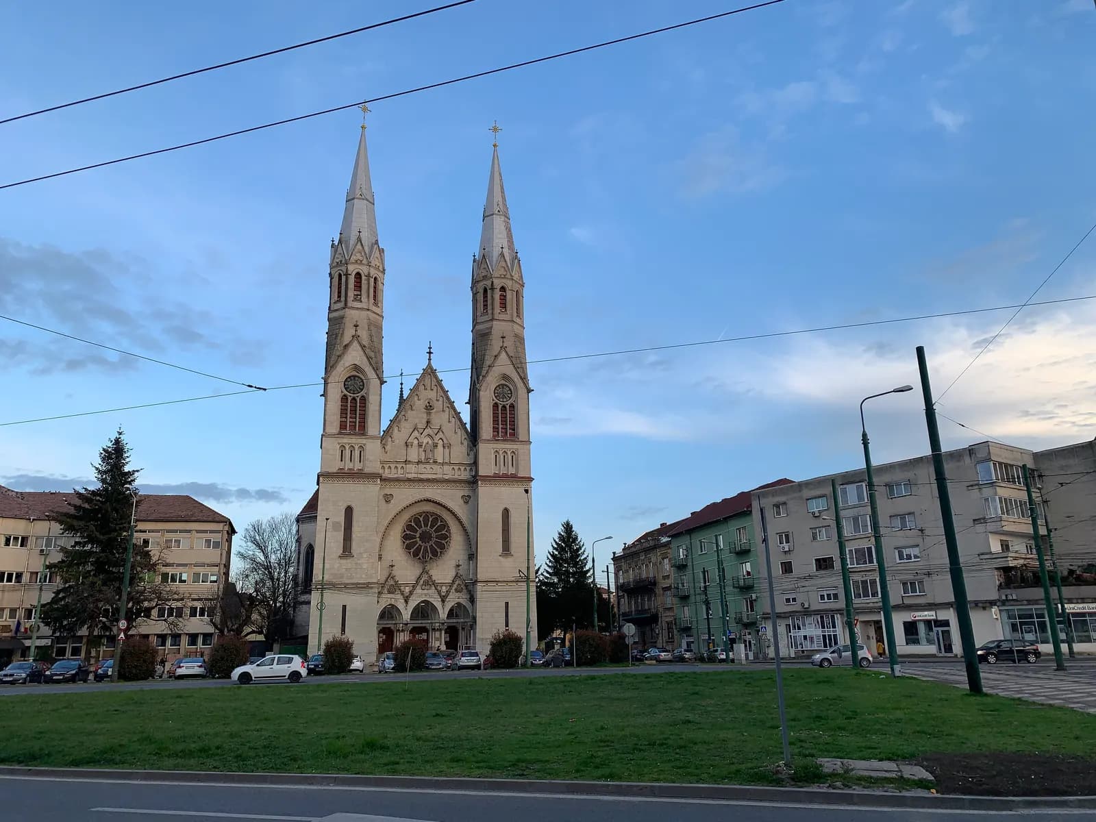 The twin-spired Roman Catholic church of Elisabetin in Timișoara, a pale neo-Gothic church with a rose window, seen across a lawn with tram wires overhead