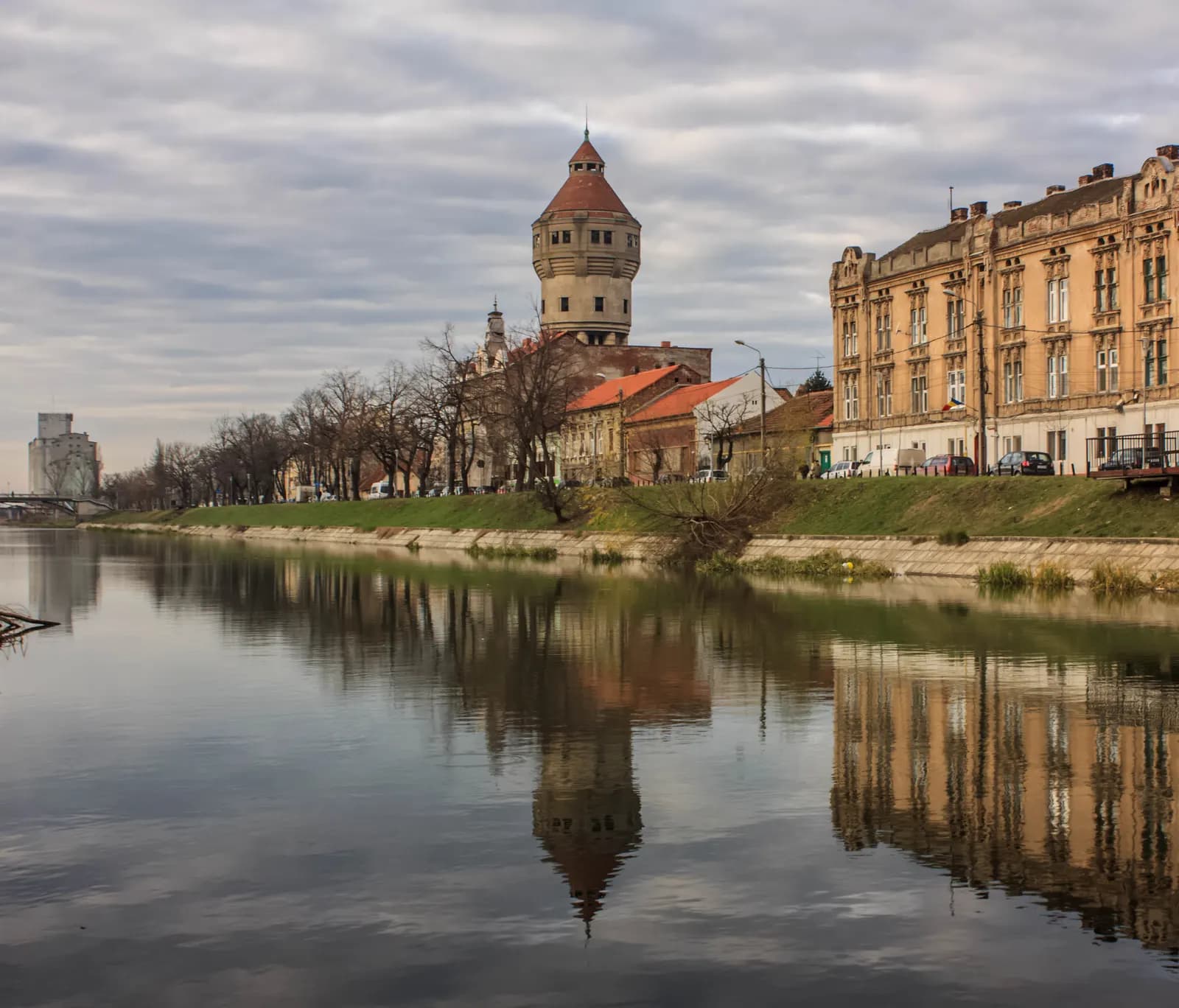 The Iosefin water tower rising behind trees and a large ochre building on the bank of the Bega canal in Timișoara, reflected in the still water
