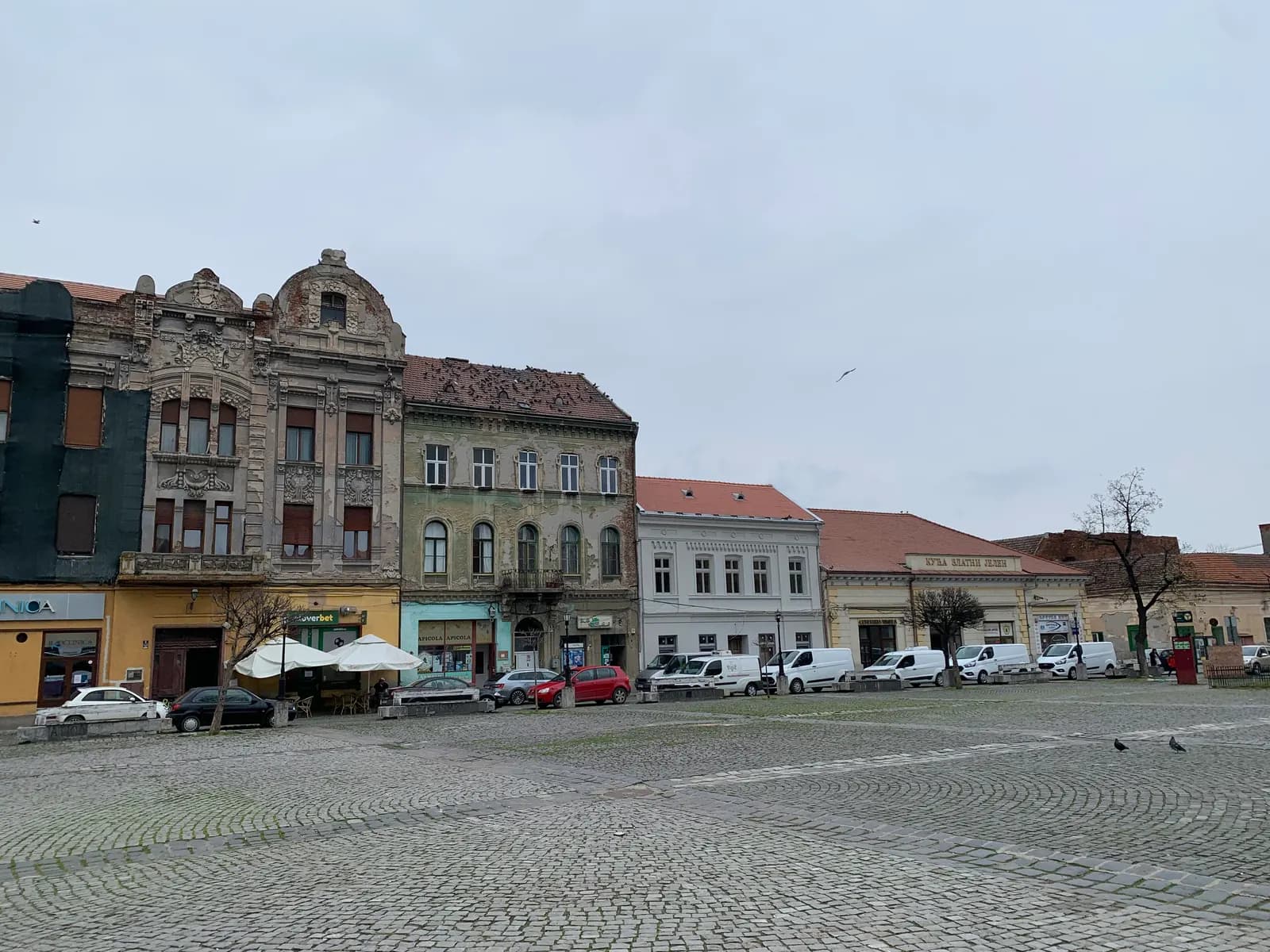 Piața Traian in Timișoara's Fabric district, a wide cobbled square edged by old two and three-storey houses with ornate facades, under an overcast sky