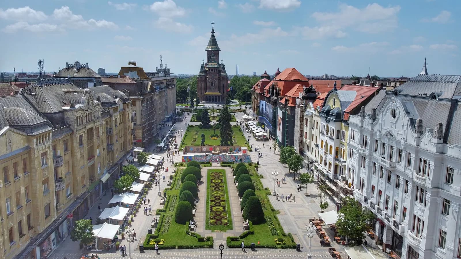 Piața Victoriei in Timișoara seen from above, a long pedestrian square with flower beds and clipped trees between rows of ornate buildings, leading to the Orthodox Metropolitan Cathedral