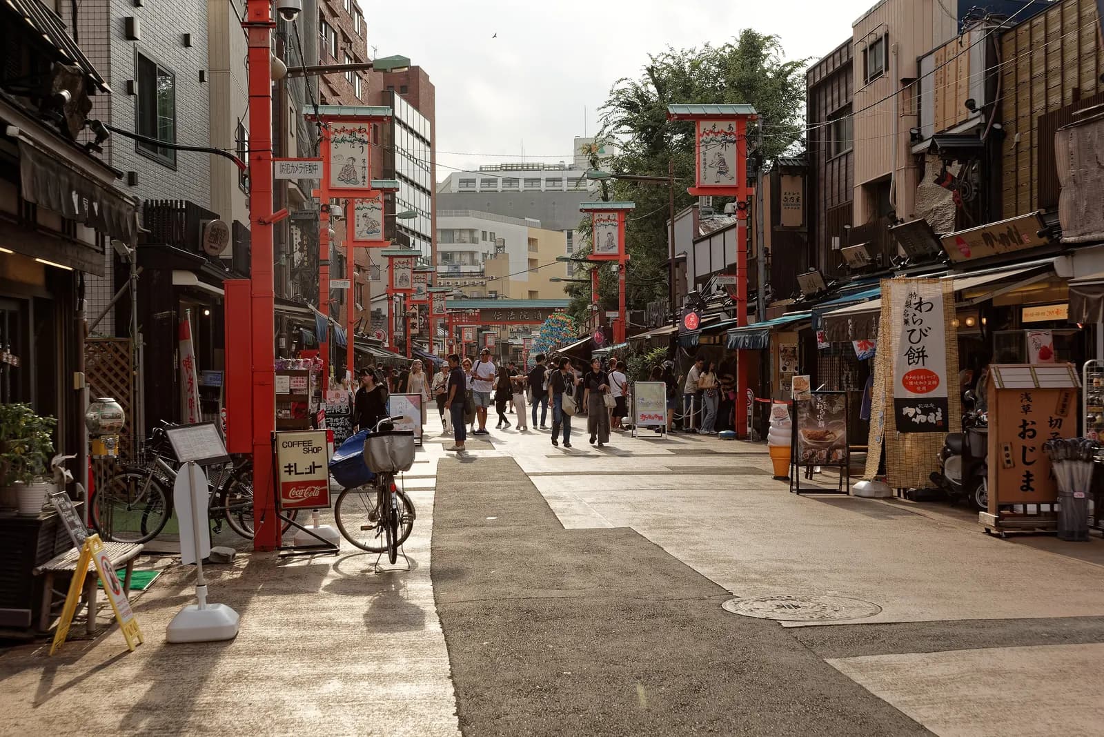 A pedestrian shopping street in Asakusa with red lantern posts, small shops and food stalls, parked bicycles and people walking in afternoon sun