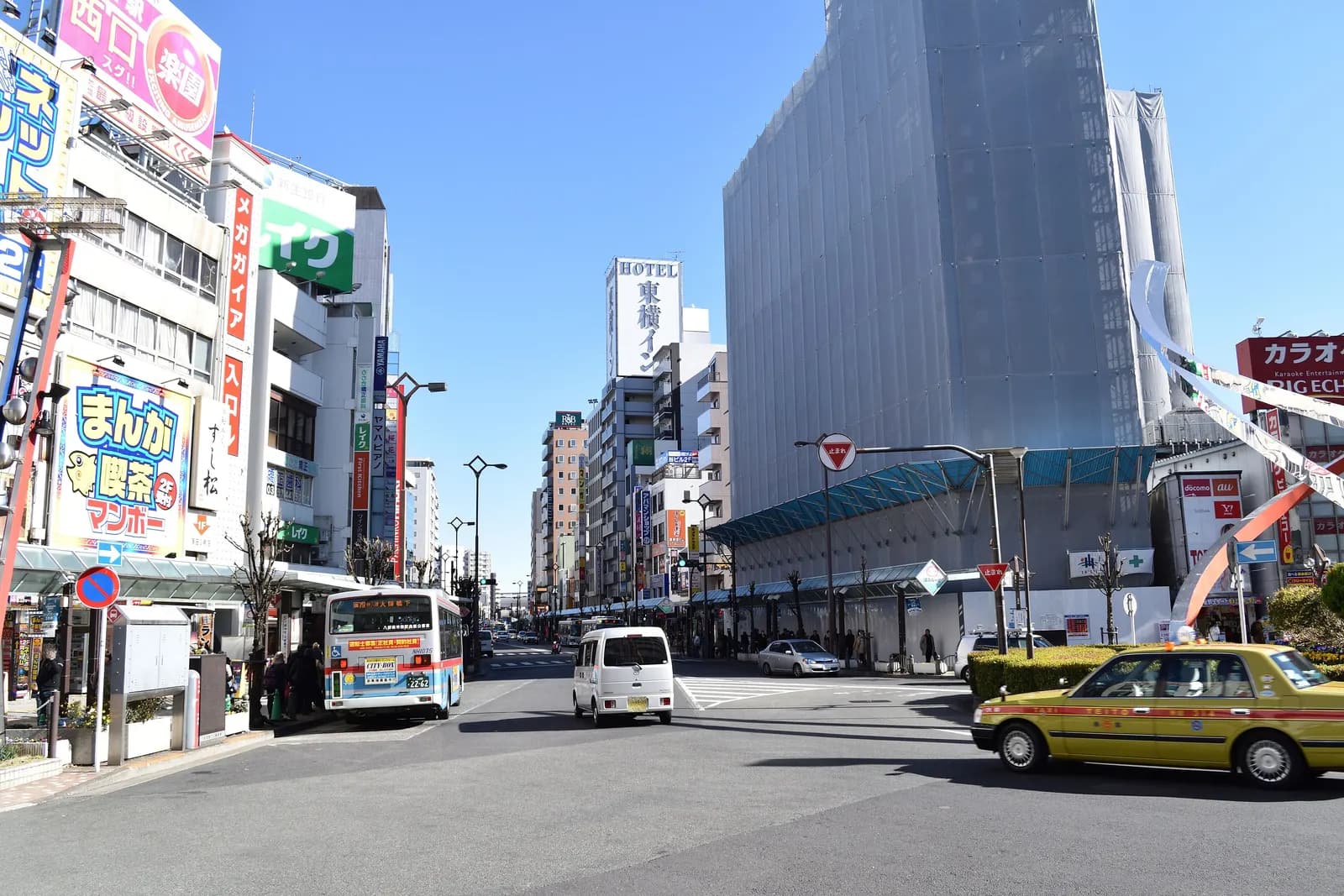 The street outside the east exit of Kamata Station, with a city bus, a taxi, shop signs and a business hotel tower under a clear winter sky