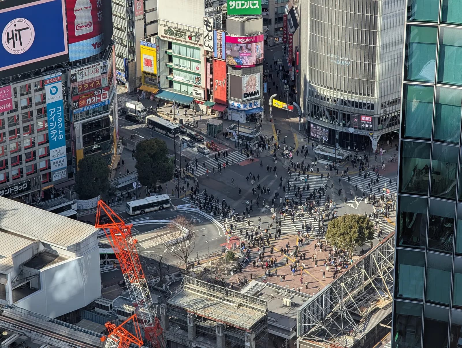 Shibuya Crossing seen from above on a winter afternoon, with people on the zebra crossings, buses on the road and a construction crane in the foreground
