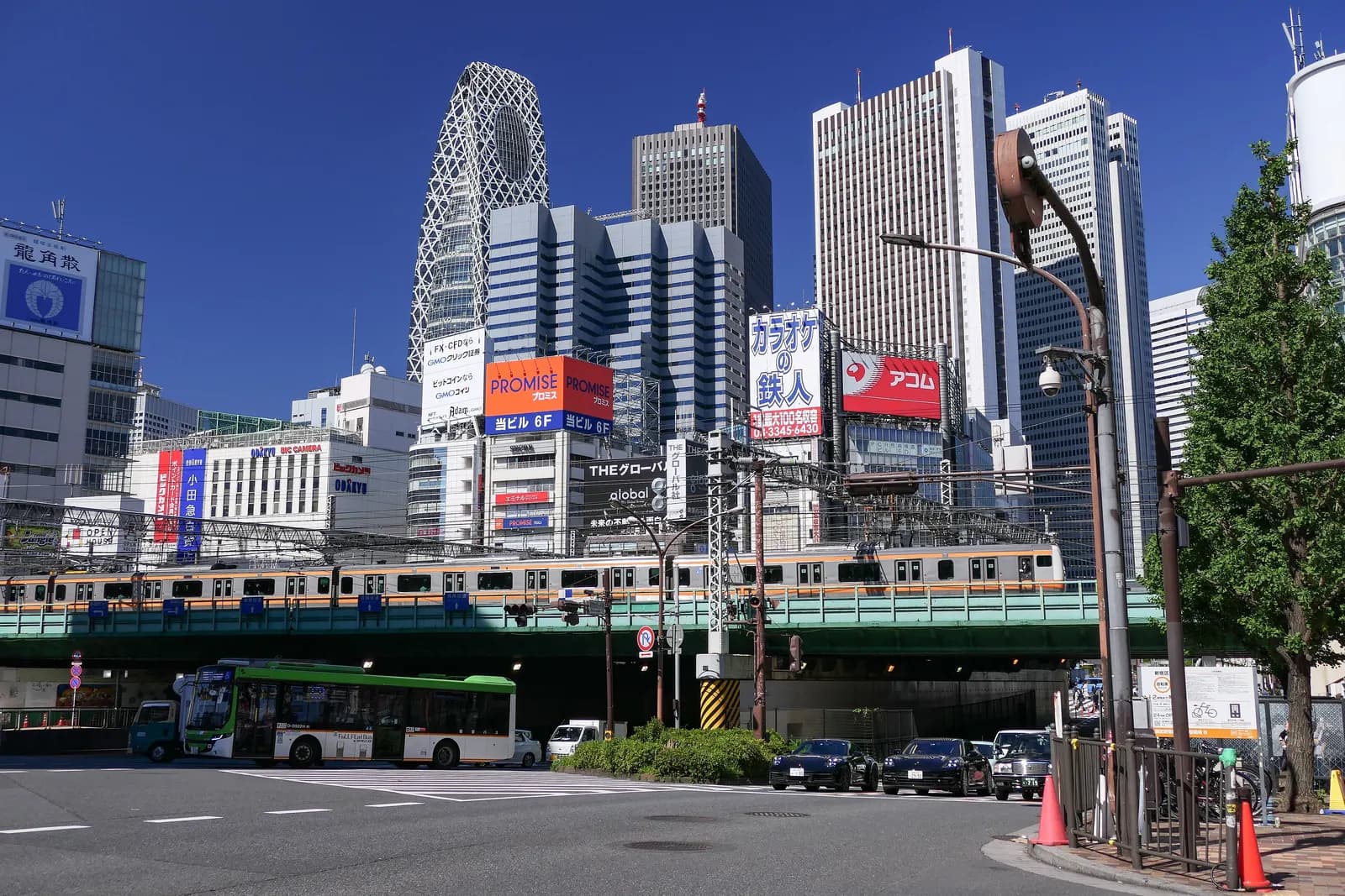 A commuter train crossing the railway bridge by Shinjuku Station, with a city bus and cars below and the Nishi-Shinjuku towers behind under a blue sky