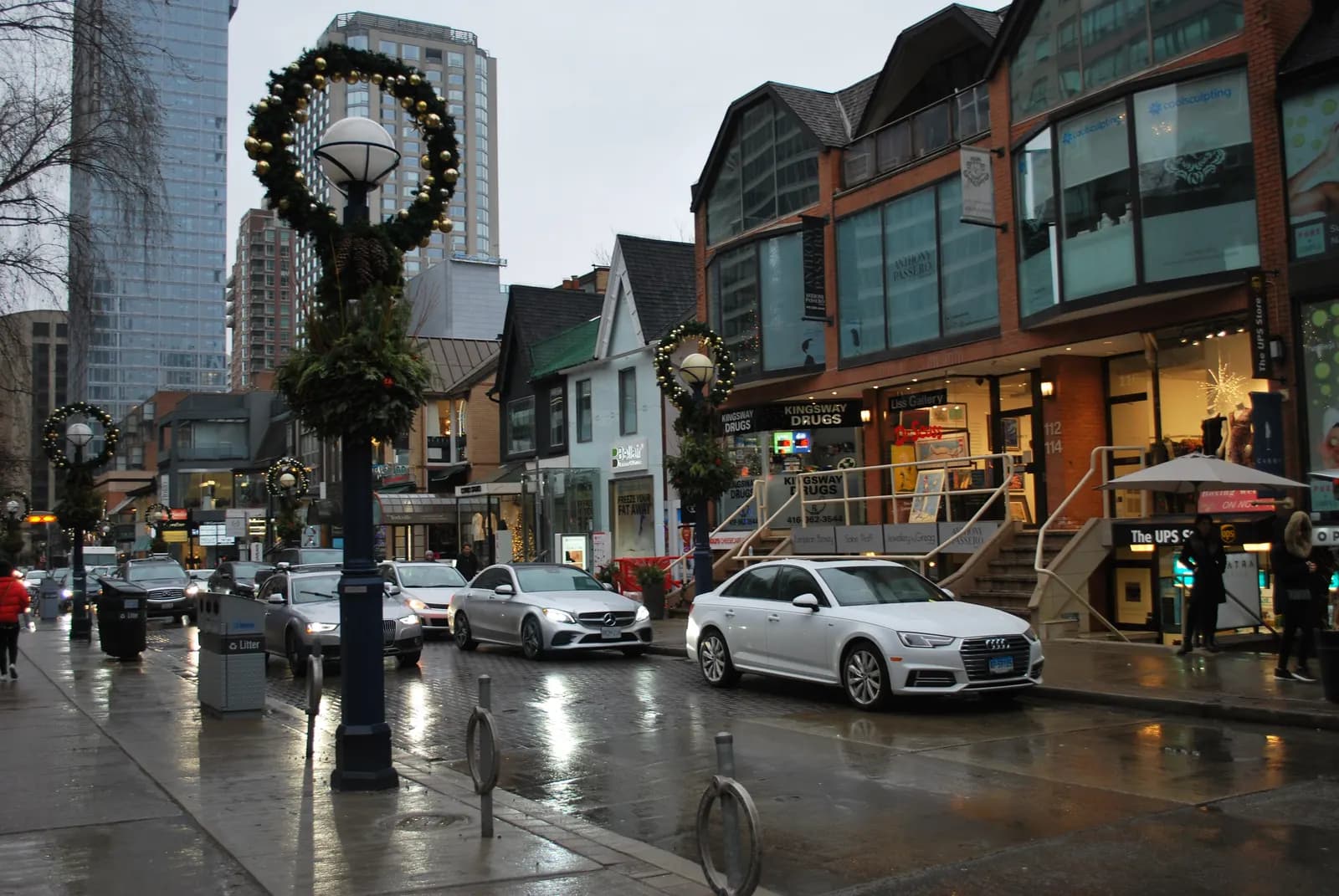 Low-rise shops and wreath-hung lamp posts on a wet Cumberland Street in Yorkville, with parked cars and towers behind