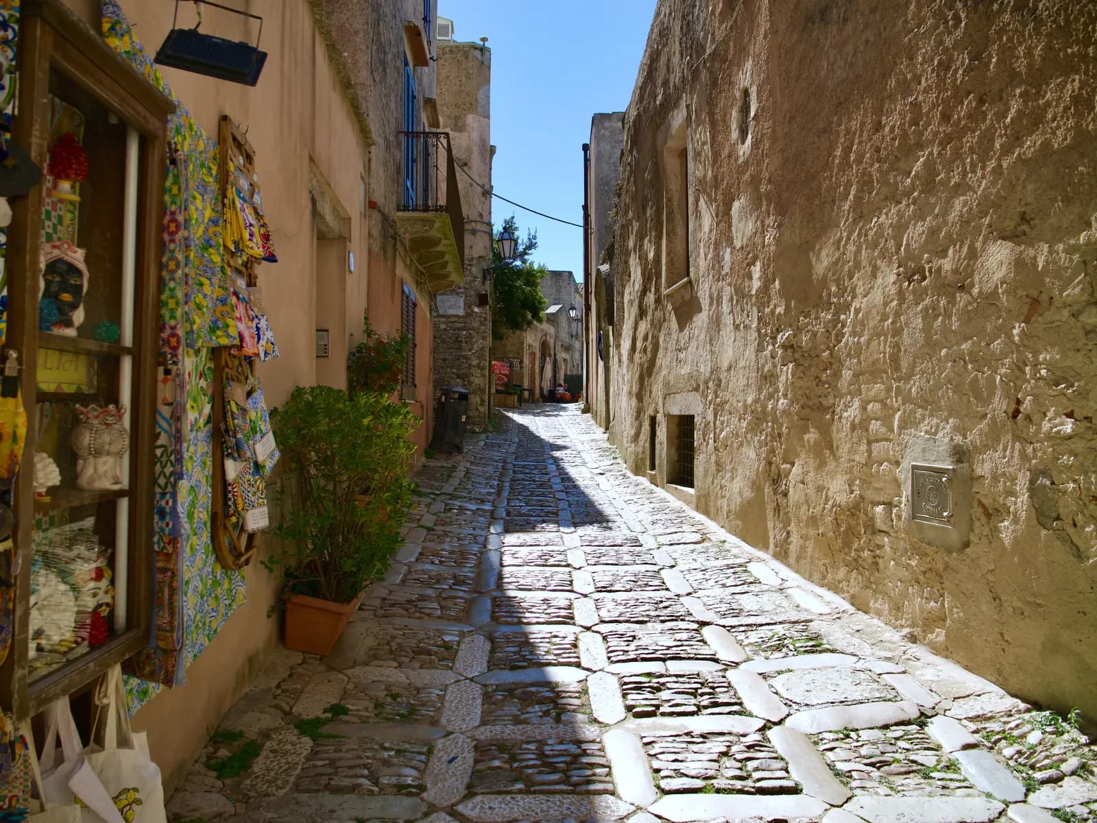 A narrow cobbled street in Erice between stone houses, with a shop display of painted ceramics on the left