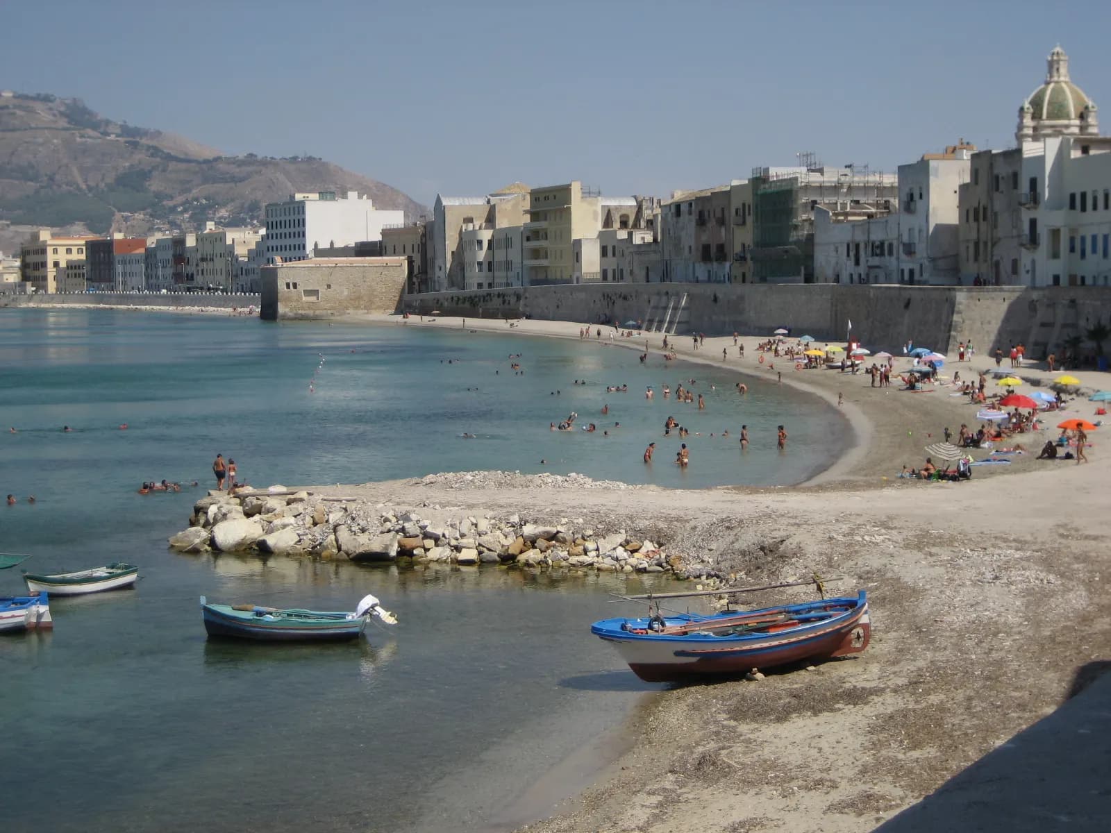 The Tramontana beach below the sea walls of Trapani, with painted boats on the sand and bathers in the shallow water