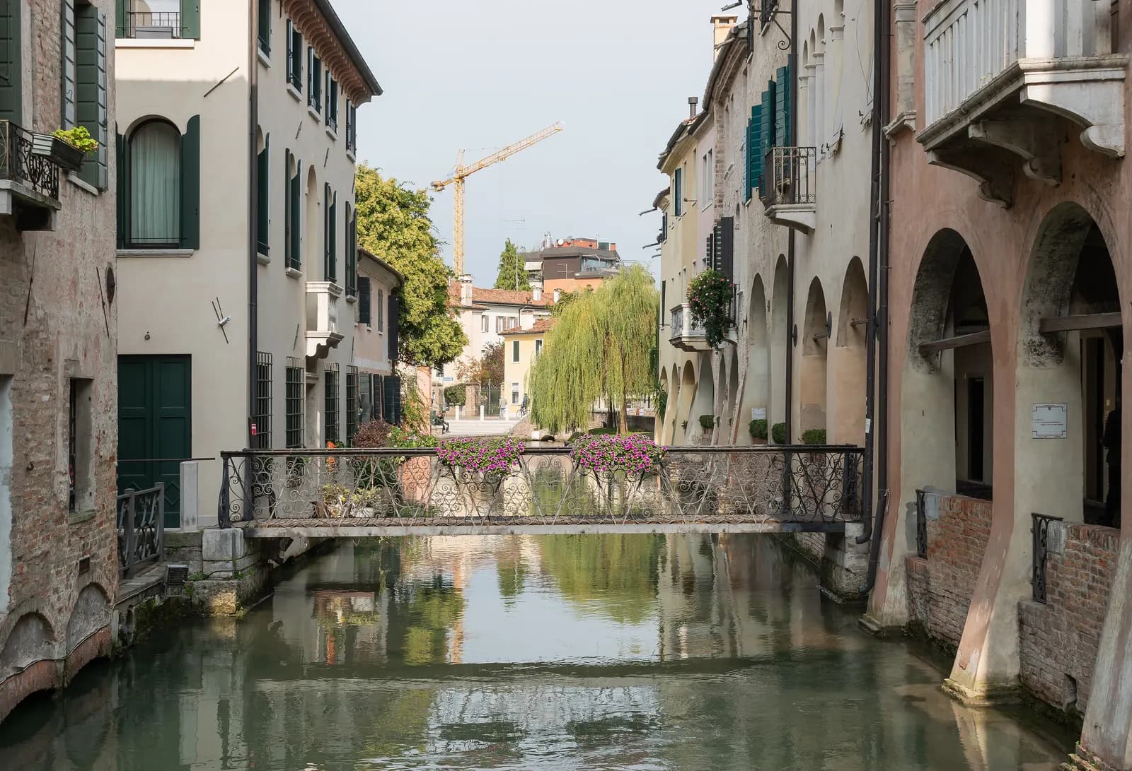 The Canale dei Buranelli in Treviso: pastel houses and an arcade along a green canal, with a wrought-iron footbridge banked with pink flowers.